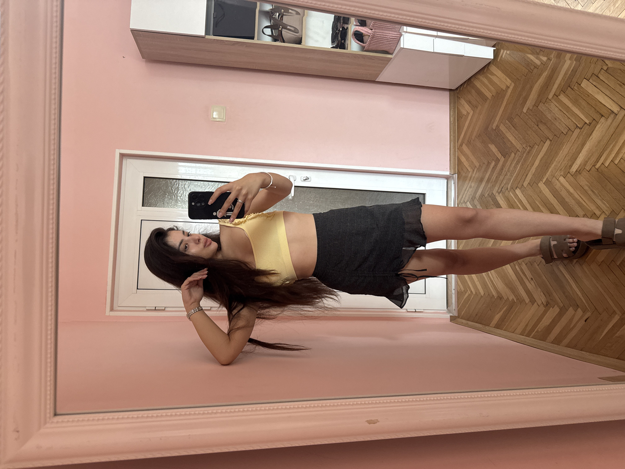 The media file displays a mirror selfie taken by a young woman in an indoor setting. The image is presented rotated 90 degrees clockwise. The woman, appearing to be in her twenties, is reflected in a large, ornate pink-framed mirror. She holds a black smartphone in her left hand, positioned to capture her reflection. She has long, dark hair styled down, partially covering her left shoulder. Her attire consists of a light yellow ribbed crop top, a dark gray or black mini skirt with ruffled drawstring details, and light brown flat sandals. She wears a silver ring on her right ring finger, a silver-toned watch, and a bracelet on her left wrist.
The room features pink-painted walls and a light-colored wooden parquet floor laid in a herringbone pattern. Above the woman's reflection, a wall-mounted cabinet with light wood and white sections is visible, containing several accessories like bags and shoes on its shelves. A white-framed window is located on the wall behind her, and a white light switch is visible on the pink wall. The scene suggests a casual moment, possibly in a bedroom or dressing area in Plovdiv, Bulgaria, during daytime. No discernible text is visible.