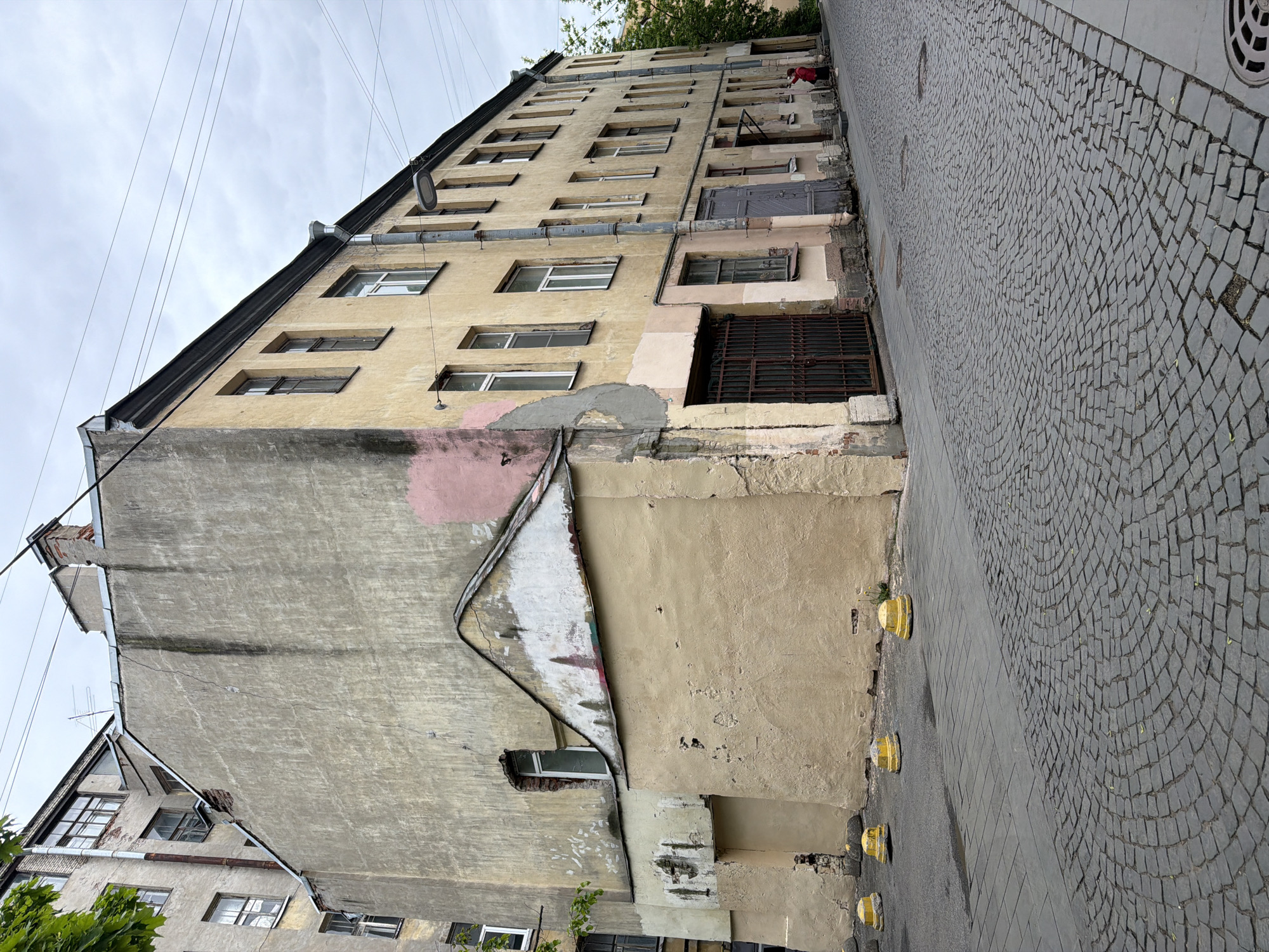 The image shows an exterior view of a building in Saint Petersburg, Russia. The building has a weathered, multi-story facade with a mix of yellow and gray plaster. Several windows are visible, some with white frames and others appearing older or with different coverings. A large, gray wall is visible on one side, showing signs of aging and repairs. The street is paved with cobblestones and concrete, and yellow bollards line the edge of the sidewalk. Overhead, there are several electrical wires.