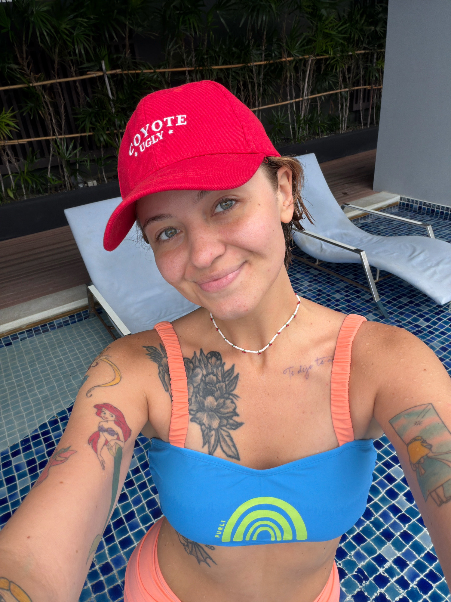 The image shows a young woman wearing a red baseball cap with white text that reads "COYOTE UGLY" with stars. She is also wearing a blue bikini top with a lime green rainbow design and coral pink bikini bottoms. She has several tattoos on her arms and chest, including a depiction of Ariel from "The Little Mermaid," a large floral design, and script writing. The woman is smiling and looking at the camera. She is in a swimming pool with blue tiled walls. In the background, there are lounge chairs and lush green foliage, suggesting an outdoor setting. The lighting indicates it is daytime, possibly late afternoon. The context of the image, including the swimwear and pool, suggests leisure or vacation. The specific location of Hua Hin City Municipality, Thailand, is implied by the provided context, indicating a tropical or subtropical environment.
