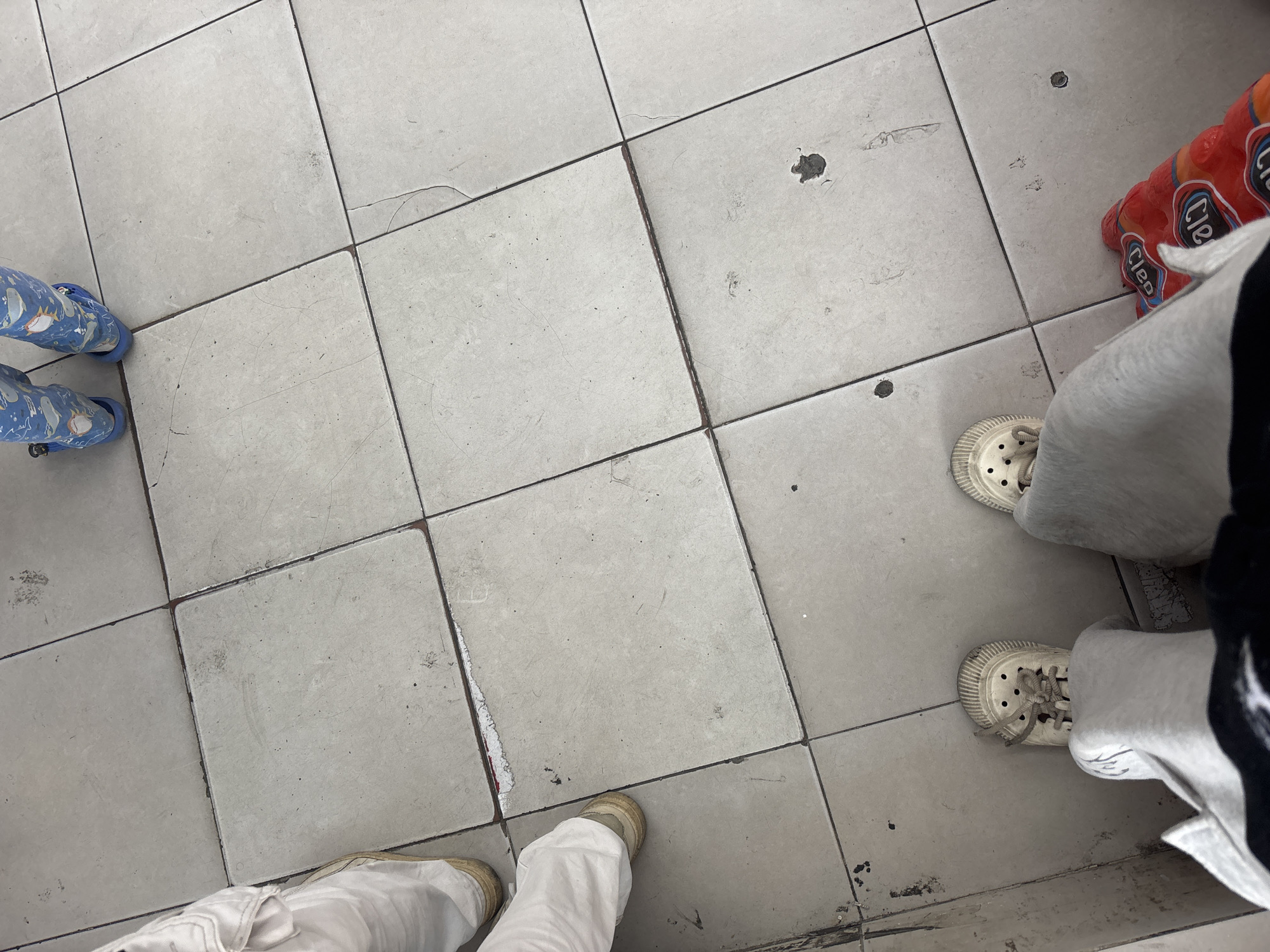 The image shows a tiled floor, likely in a public indoor space, with several pairs of feet visible. In the lower part of the frame, a person's feet are clad in white, hole-covered shoes and light gray pants. To the right of these feet, a red package of what appears to be toilet paper is partially visible. Further up and to the left, another person's legs and feet are seen, wearing light-colored pants and tan shoes. At the top of the image, two small legs are visible, wearing blue pajamas with a whale pattern and blue sandals. The tiles are light gray and square, with visible grout lines and some wear and tear, including cracks and dark smudges. The scene appears to be in North Jakarta, Indonesia, possibly a retail or public restroom area due to the presence of toilet paper and the tiled flooring.