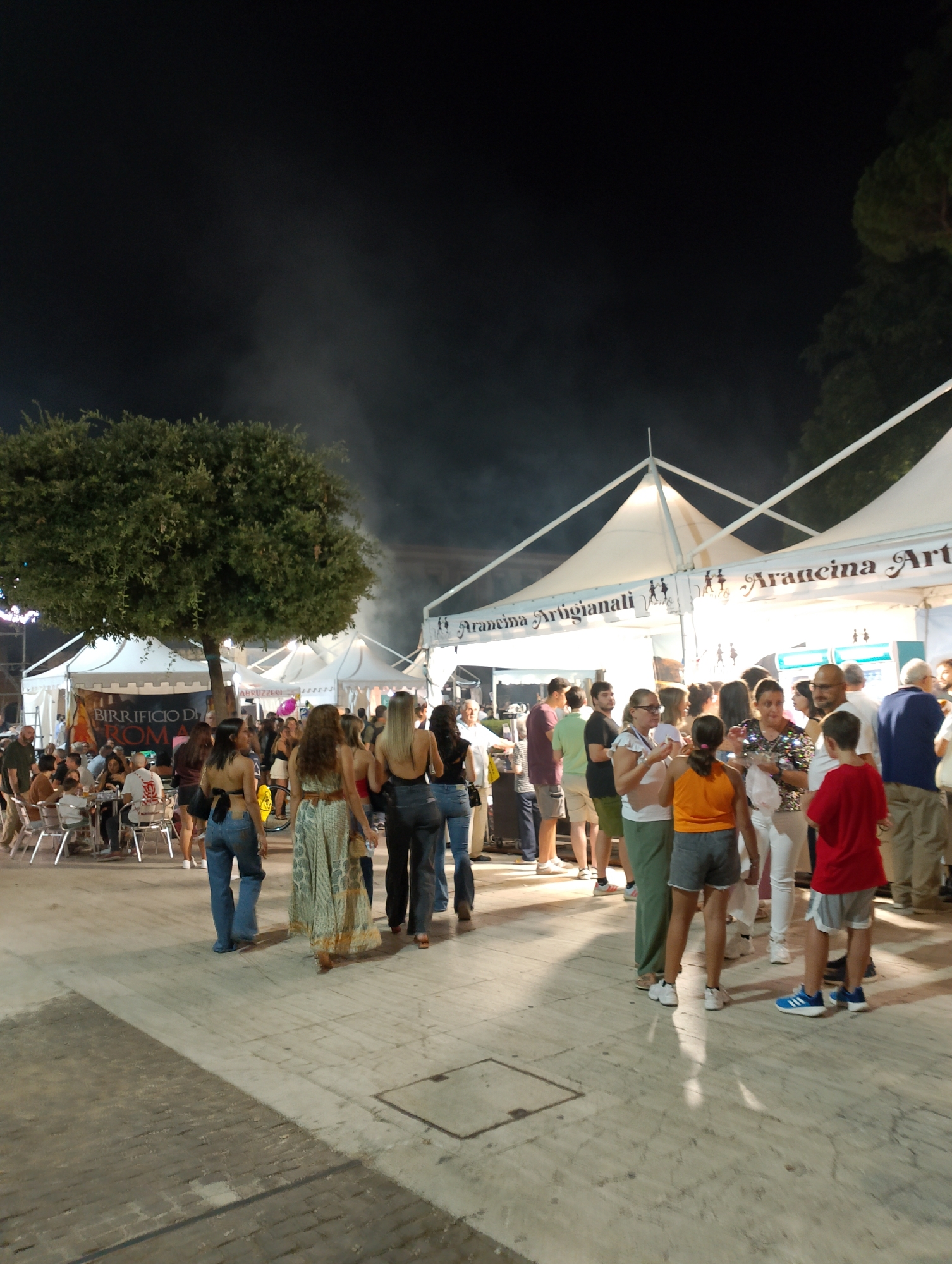 The image depicts a night scene at an outdoor event, possibly a festival or market, in Rieti, Italy. Several white tents with triangular roofs are set up, some displaying text like "Birrificio di Roma" and "Arancina Artigianali." A large tree with dense foliage stands prominently in the left half of the frame. A crowd of people, dressed in casual summer attire, are milling around, socializing, and browsing the stalls. Some individuals are seated at tables, while others are standing and talking. The lighting suggests artificial illumination from the tents and possibly streetlights. The ground appears to be paved. The overall atmosphere seems lively and social.