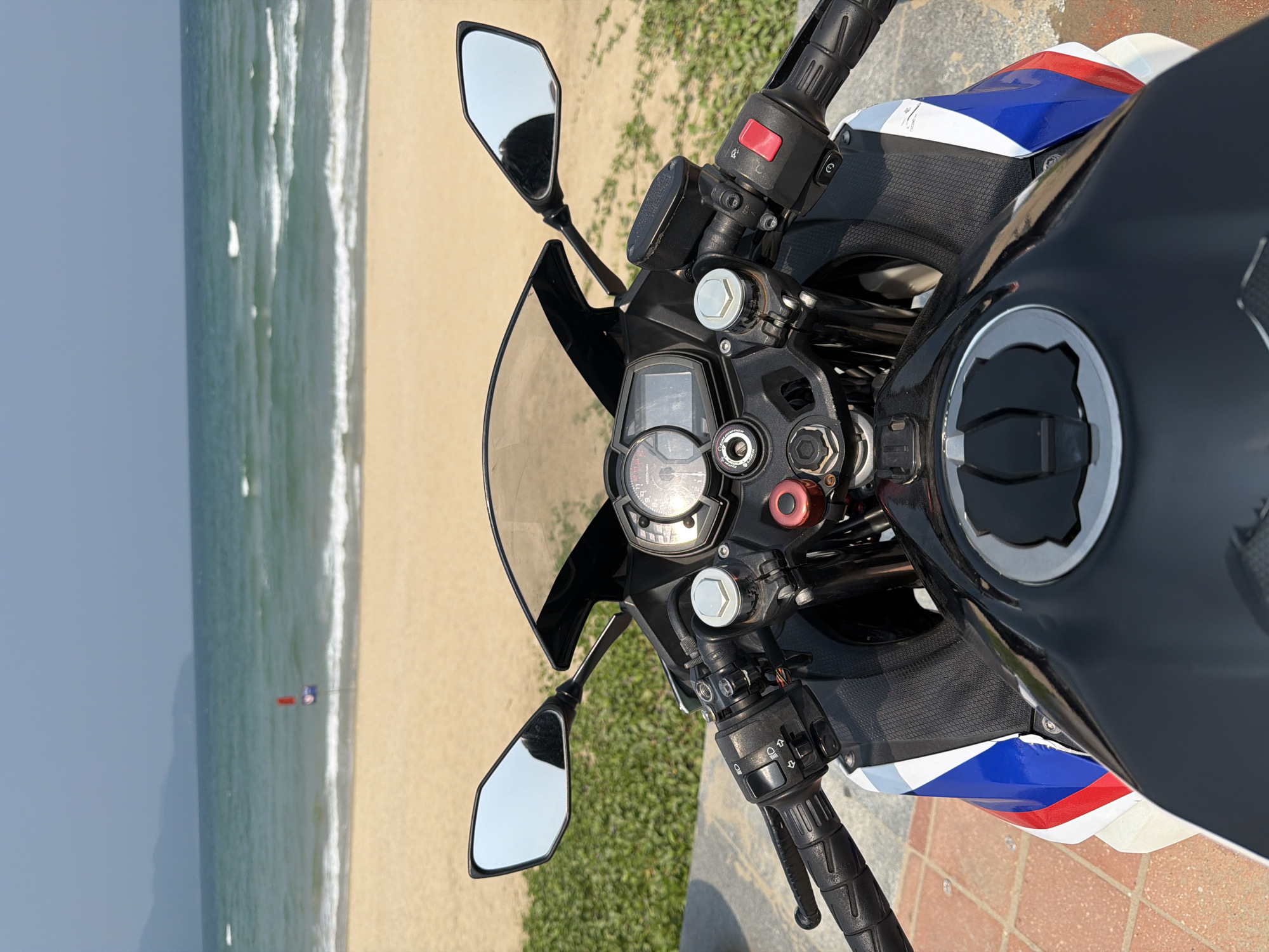 From an elevated point of view, the image captures the dashboard and handlebars of a motorcycle. The motorcycle is parked on a brick-paved surface, next to a patch of green foliage. In the background, there is a view of a beach with gentle waves meeting the shore. Further in the distance, there's an expanse of water and a slightly hazy sky. The beach is likely My Khe Beach in Đà Nẵng, Vietnam. A red flag is seen on the beach near the water.