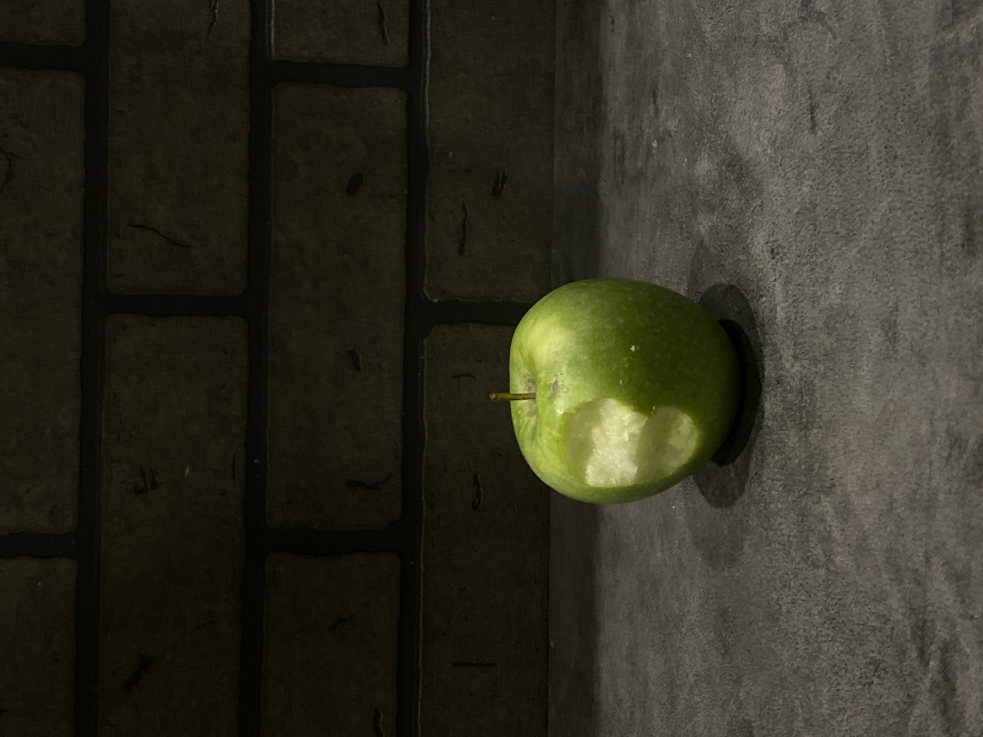 A half-eaten green apple is positioned on a dark gray wall in the corner of a room. The wall to the left is a brick pattern. The lighting is dim and creates shadows. The scene appears to be indoors. No people are visible. There is no indication of the location being Sochi, Russia.