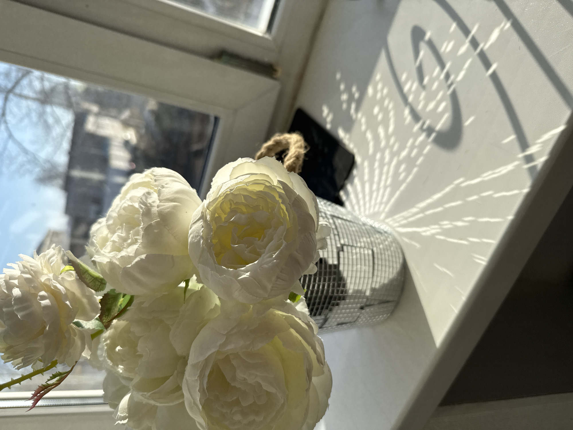 A close-up, high-angle shot shows a bouquet of artificial white peonies in a reflective, mirror-tiled vase on a white windowsill. Reflected sunlight creates patterns on the windowsill. Through the window, a blurry, indistinct cityscape is visible, consistent with an urban setting, which could be Chaikovsky, Russia. A dark object, possibly electronic, sits partially obscured behind the vase. There are no people visible in the image.