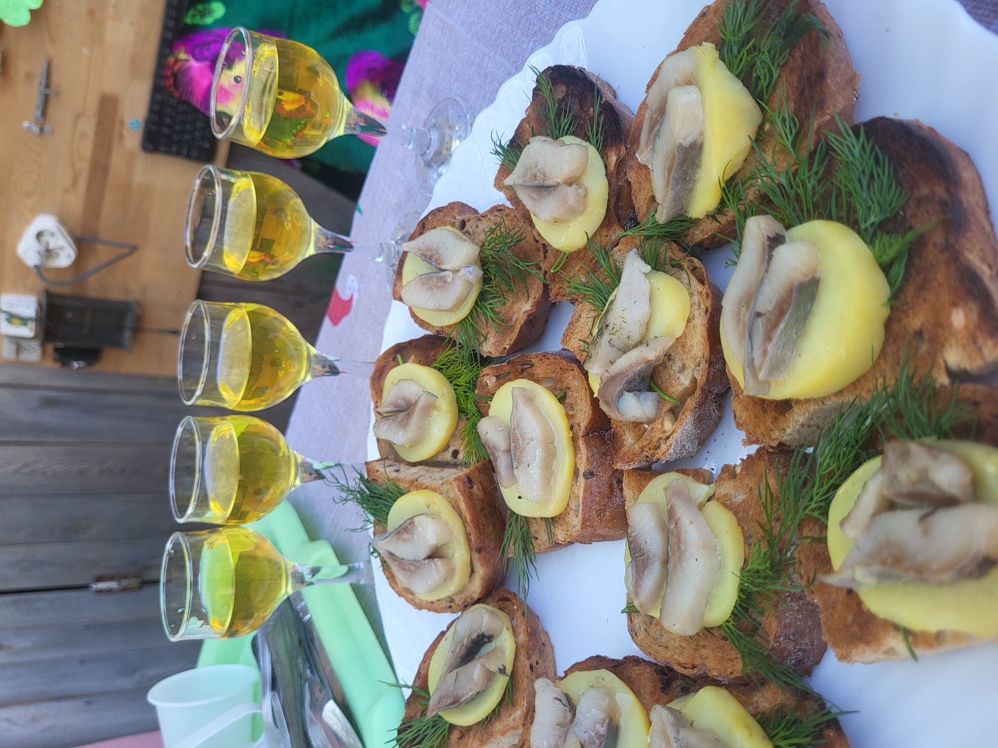 Here is a detailed description of the image:
**Scene:** A table setting, possibly outdoors, with an arrangement of appetizers and drinks.
**Objects:**
* A white plate is prominently featured, holding several open-faced sandwiches. The sandwiches consist of toasted bread slices topped with what appear to be slices of potato, pieces of herring, and sprigs of dill.
* To the left, a row of wine glasses filled with a yellow liquid (likely white wine or a similar beverage) are lined up.
* Other items such as a green tablecloth or napkin, utensils (possibly forks or spoons), and a white cup are visible in the background.
**Possible Location (Based on prompt):** Given the prompt specifies Зипуново, Russia, the setting is likely a gathering or meal taking place in this city. The background suggests an outdoor location.
**Actions/Interactions:** The image suggests a prepared meal or appetizer selection ready to be consumed. No people are directly visible, but the setup indicates an event where eating and drinking are likely taking place.