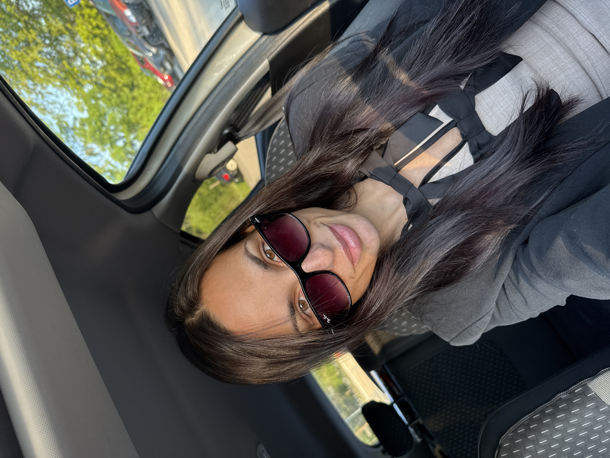 A woman with long brown hair, wearing sunglasses, a gray top with a bow, and a black jacket, is sitting in a car. The car is stationary, as indicated by the blurred trees and other vehicles visible through the window. The woman is looking directly at the camera with a slight smile. The location is not discernible from the image to be specifically in Pleven, Bulgaria. The scene is well-lit, suggesting daylight hours.