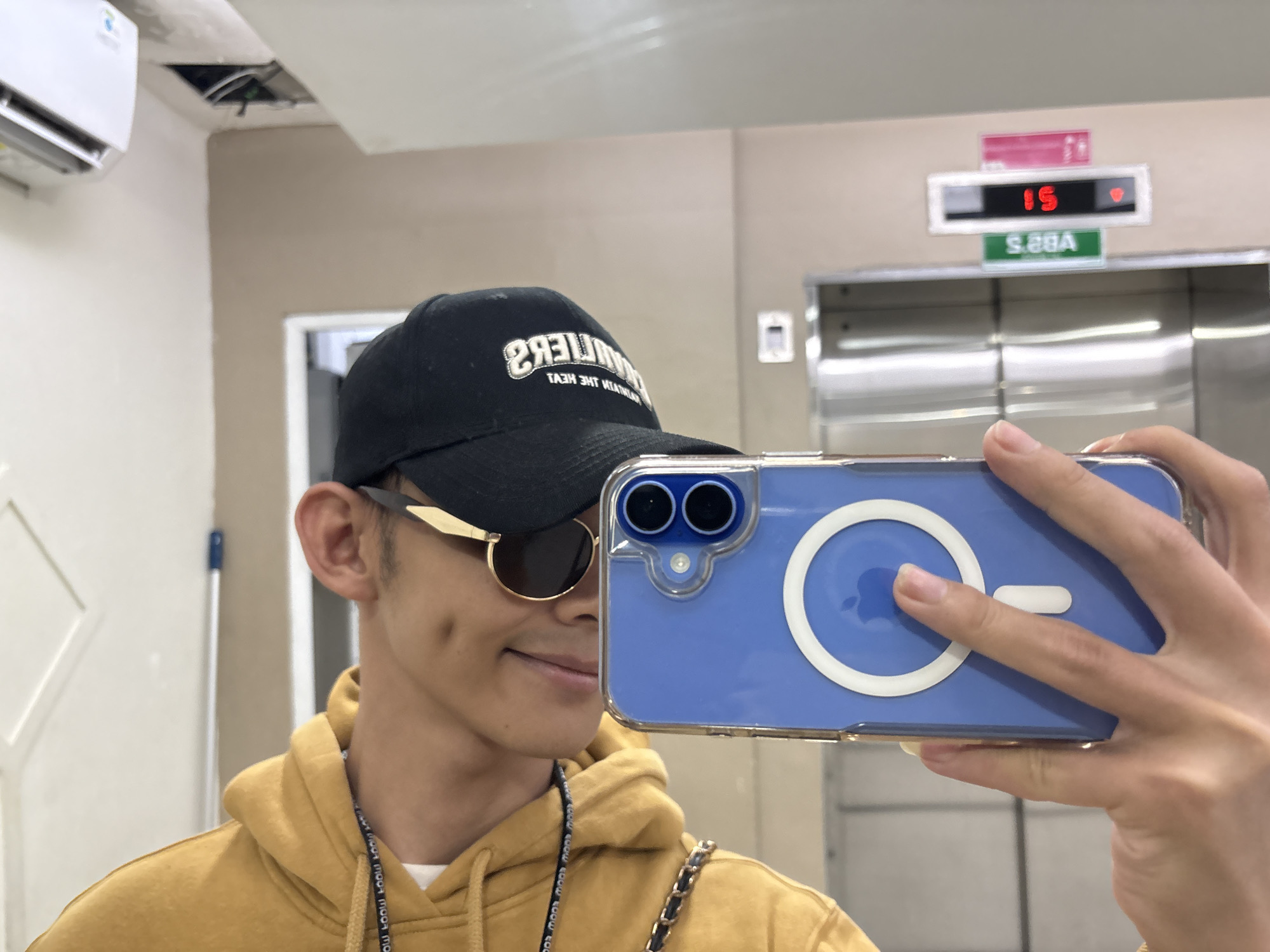 A young man with dimples and dark hair is taking a selfie in what appears to be an elevator lobby. He is wearing a black baseball cap, sunglasses with gold rims, and a mustard yellow hoodie. The cap has white text that is difficult to read clearly in the reflection. Around his neck, a lanyard with repeating black text is visible. In his right hand, he is holding a light blue iPhone with a clear case that reveals a white circle on the back.
The background shows a wall with a section of exposed wiring in the ceiling, suggesting possible ongoing construction or maintenance. To the left, a white air conditioning unit is mounted high on the wall. To the right, an elevator door with a brushed metal finish is visible, along with an illuminated display above it. The display shows "15" in red digits, indicating the 15th floor, and below it, a green sign with white text reads "S.GBA".
The lighting suggests it could be daytime, but the indoor setting makes it difficult to determine. The man has a slight smile, and the overall mood of the photo seems casual and self-assured. The presence of the elevator and the building's interior suggests an urban environment, and the prompt specifies the location as North Jakarta, Indonesia.