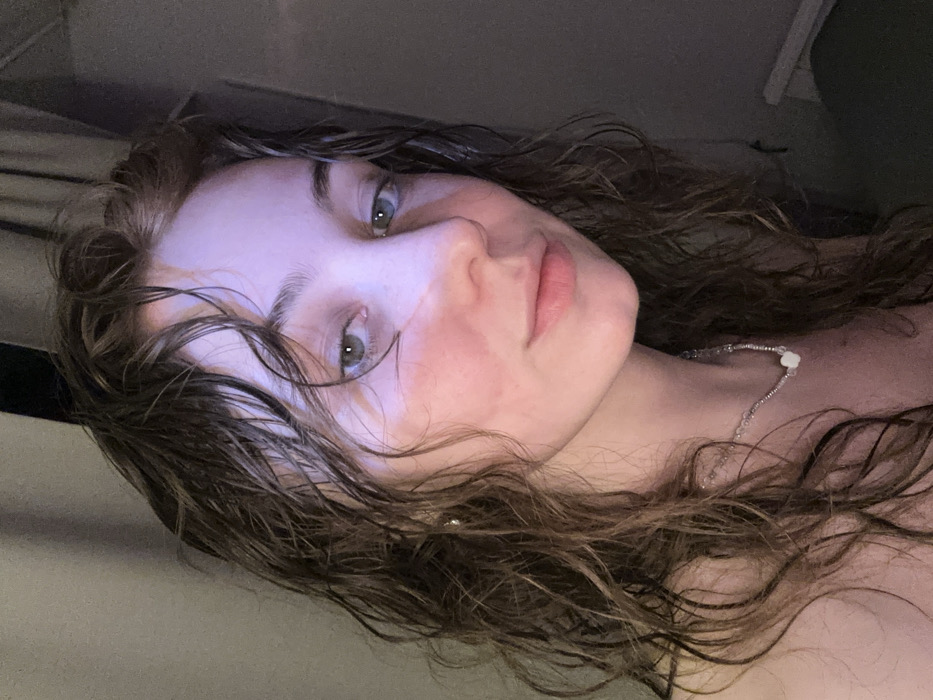 A single female individual is depicted in a close-up, chest-up portrait. She possesses long, wet, dark brown, wavy hair. Her eyes are light, appearing greenish-blue. A distinct lighter band, possibly a tan line or light reflection, spans horizontally across the bridge of her nose and upper cheeks. Her lower cheeks show a reddish tint. She wears a delicate necklace composed of small, clear beads and a central white, possibly floral-shaped, bead. Her shoulders appear bare. The individual's gaze is directed forward towards the camera, and her lips form a slight, closed-mouth smile. The background is dark, out of focus, and indistinct, suggesting an indoor setting with low ambient light. A vertical dark line is visible on the far right, consistent with a curtain or wall edge. A cool-toned, possibly blue or purple, light source illuminates the left side of her face and hair, contrasting with warmer tones elsewhere. No visual elements within the image provide specific information to identify the location as Moscow, Russia.