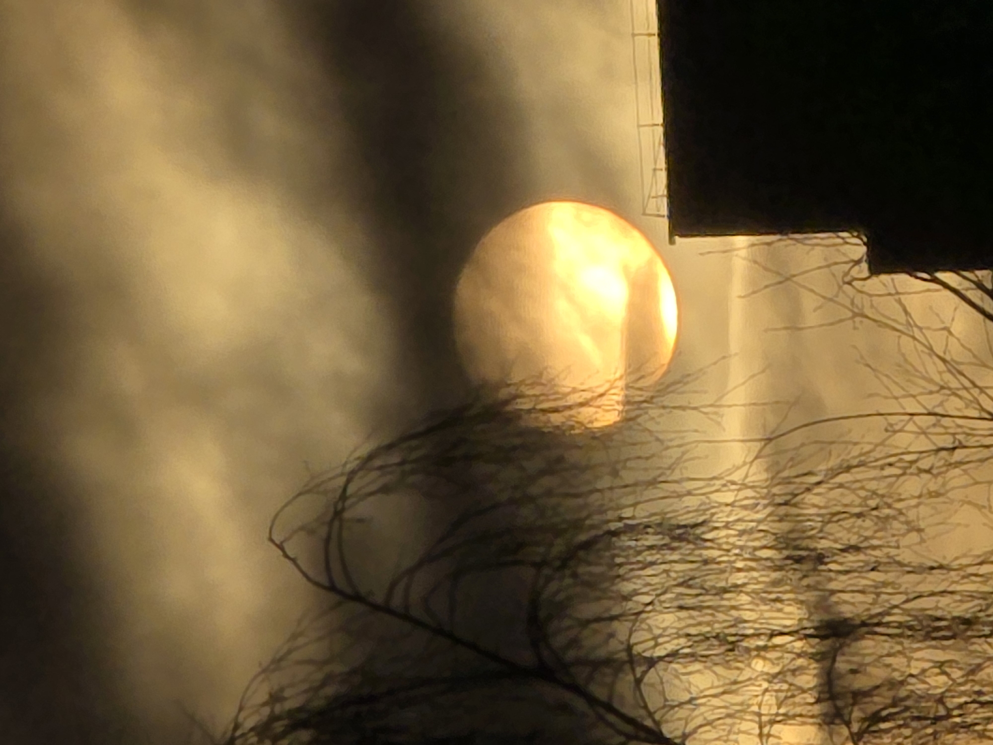 The image shows a hazy, golden-lit scene. A bright, round object resembling the sun is obscured by clouds. Silhouetted tree branches are visible in the foreground. The upper right corner contains a dark, indistinct structure, possibly a building with a ladder. The location is Unknown, Unknown. There are no people visible in the image. The environment is overcast.