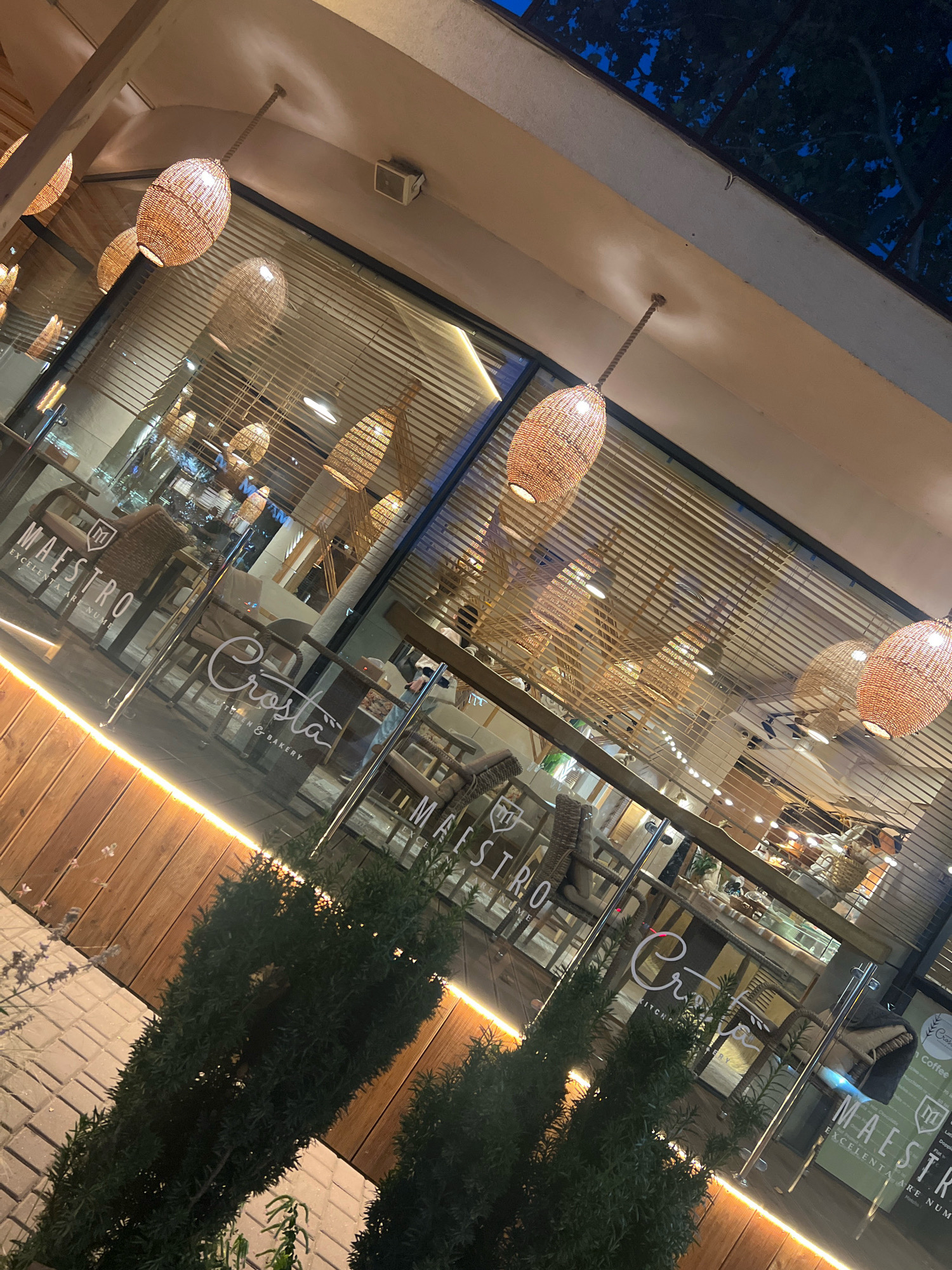 A nighttime exterior view of a restaurant or cafe with large glass windows and doors. The establishment has a warm, inviting ambiance, with numerous woven pendant lights illuminating the interior and reflecting on the glass. Inside, wicker chairs and wooden tables are visible, suggesting a cozy and rustic decor. The exterior features wooden paneling along the base and a bright strip of light accentuating the architectural lines. Green bushes are planted in front of the building, adding a touch of nature. The reflection of the lights and trees in the glass creates a layered visual effect. The signage on the glass indicates the names "MAESTRO" and "Crosta KITCHEN & BAKERY". The overall scene is clean and well-maintained, conveying a sense of a popular dining or gathering spot.