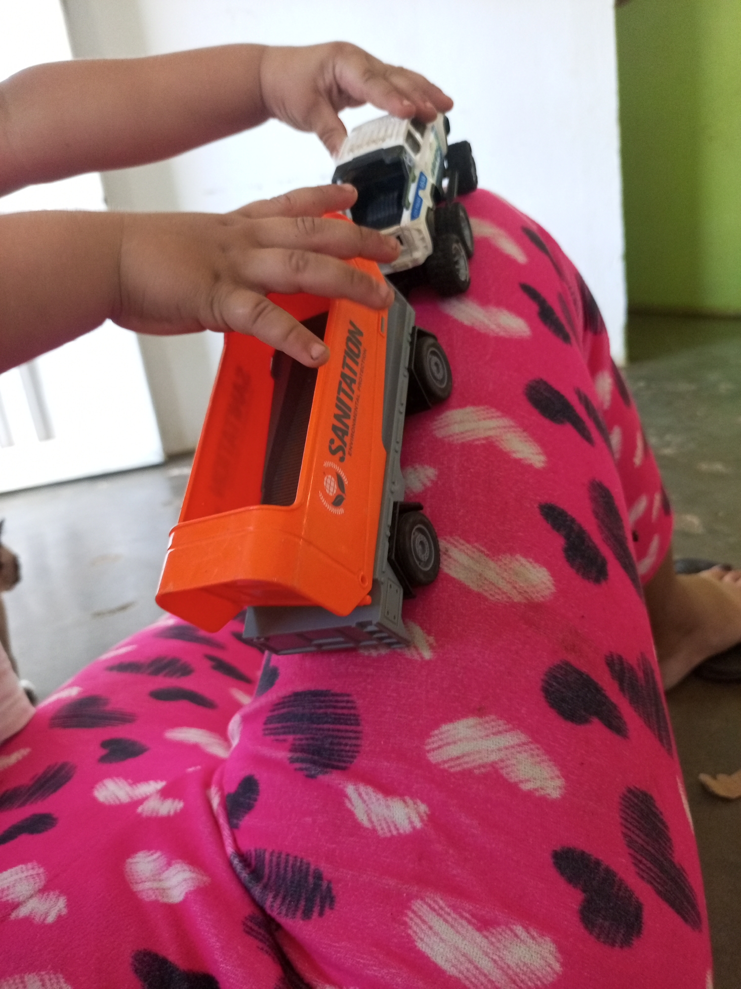 In an indoor setting in La Tagua, Venezuela, a close-up shot focuses on the hands of a child playing with a toy sanitation truck. The truck is orange and grey, with "SANITATION ENVIRONMENTAL PROTECTION" written on its side. The child's hands are on the toy, manipulating it. The child is wearing bright pink pants with a heart-shaped pattern in black and white. The background is blurred, showing a white wall and a hint of a green wall. The lighting suggests it is daytime.