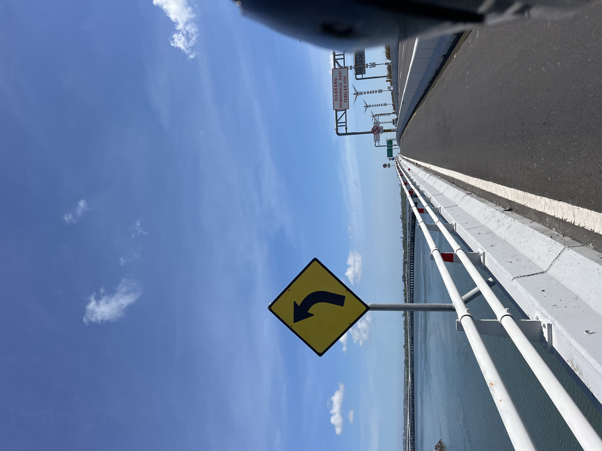 A road on a bridge over water with a bright blue sky and some clouds. A yellow diamond-shaped sign with a black arrow pointing left is on the left side of the road. There are white railings and a concrete barrier along the edge of the bridge. In the distance, there are more bridges or structures. A black object, likely part of a vehicle, is in the foreground on the right. A sign above the road in the distance has red text. This is a view from a moving vehicle, likely a motorcycle, traveling on the Jagatnatha Bridge in Denpasar, Bali, Indonesia, a prominent infrastructure project connecting the Sanur area to the island of Nusa Lembongan.