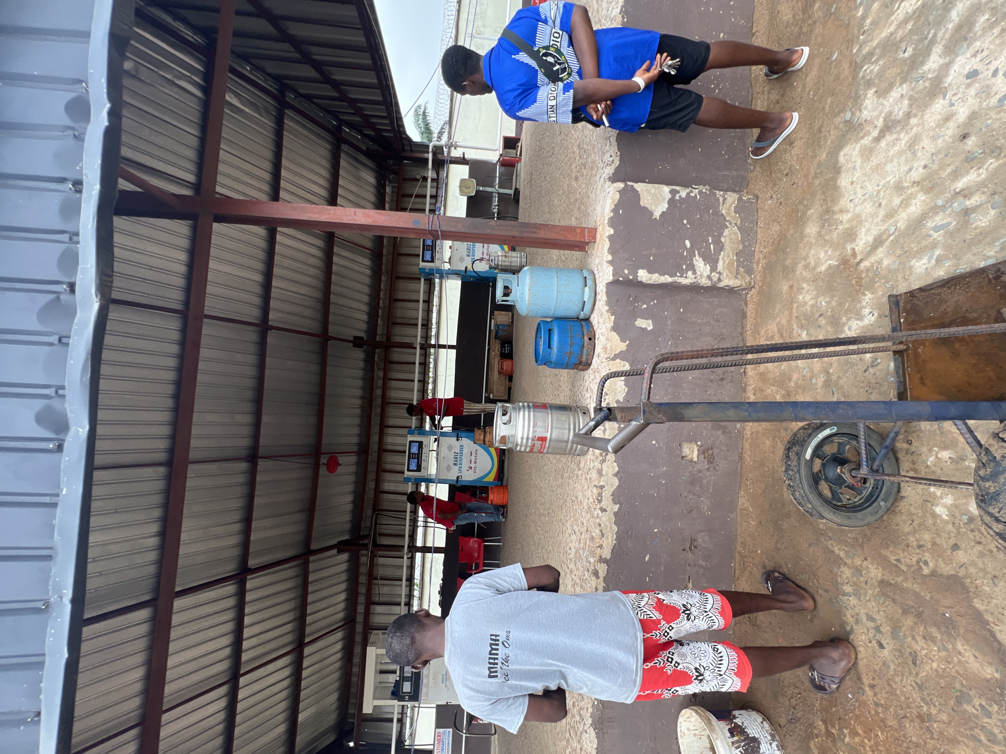 This media file depicts a scene at a Liquefied Petroleum Gas (LPG) dispensing station in Nkpor, Nigeria. Two men are the primary subjects. The man in the foreground is wearing a light grey t-shirt with the text "MAMA ee Ike Oma" on the back and red and white patterned shorts. He is standing near a cart with a wheel. The second man, in the background, is wearing a blue t-shirt with "CHRISTIAN DIOR" visible and black shorts. He is holding keys.
The setting is an outdoor covered area, likely a gas station with a metal roof supported by red metal beams. Several LPG cylinders are visible, with one large silver cylinder and two smaller blue ones in the foreground. An LPG dispenser machine, labeled "HARIZ LPG DISPENSER OIL-Metals", is also present in the background. Other individuals in red shirts can be seen working at or near the dispenser. The ground appears to be a rough concrete surface.
The activity appears to be related to the operation of the LPG station, possibly dispensing gas or managing the cylinders. The lighting suggests it is daytime, and the weather appears overcast or mildly bright. The overall mood is functional and commercial, with no strong emotional cues evident. The presence of the text on the man's shirt and the dispenser provides specific identifying details.