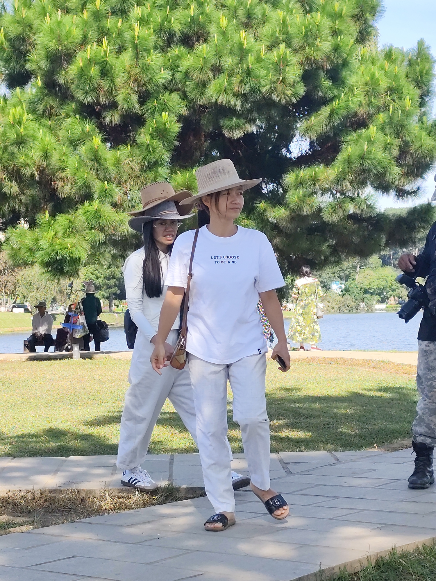The image captures a scene at Xuan Huong Lake in Da Lat, Vietnam. In the foreground, two young Vietnamese women are walking on a paved pathway. The woman on the right, facing slightly left, wears a white t-shirt with blue text that reads "LET'S CHOOSE TO BE KIND" and white pants. She is also wearing a wide-brimmed straw hat and carrying a small brown purse over her shoulder. The woman on the left is walking behind her and slightly to the left. She is wearing a white long-sleeved shirt, white pants, and a grey and brown wide-brimmed hat stacked on top of another similar hat. She is also wearing glasses. In the background, a large pine tree dominates the left side of the frame. Beyond the pathway and the tree, a body of water, presumed to be Xuan Huong Lake, is visible. On the far side of the lake, trees and some buildings can be seen. Several other people are present in the background, including a man and woman sitting on a bench near the water's edge, and another person walking away from the camera towards the lake. A person in camouflage pants and black boots, holding a camera, is visible on the far right of the frame. The overall impression is a sunny day in a park-like setting.