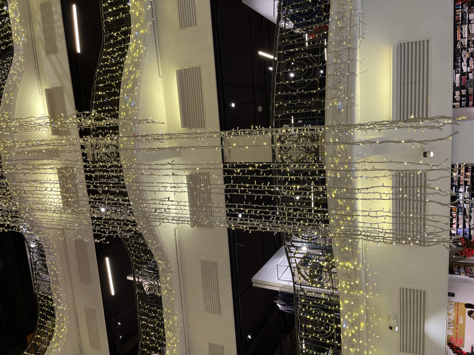 The image displays a decorated interior space, likely a shopping mall, viewed from below. Strands of small, warm white lights crisscross the ceiling, creating a grid-like pattern. The lighting is interspersed with architectural elements, including white pillars, black vertical structures, and decorative ironwork balconies. The overall impression is one of festive ornamentation and spatial complexity.