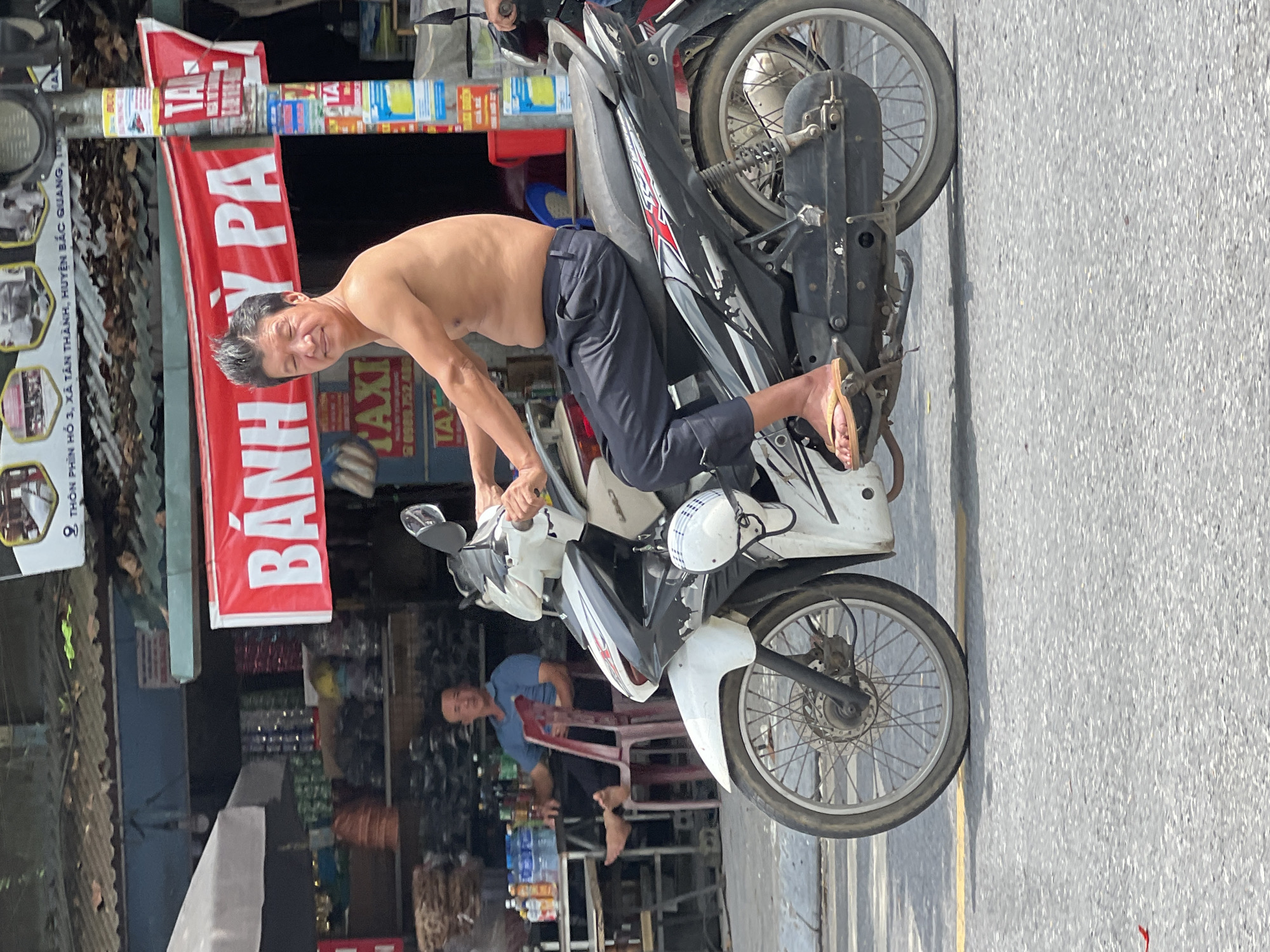 A shirtless man with slicked-back black hair rides a white and black motorcycle down a paved road in Tân Quang Commune, Vietnam. The man is wearing dark pants and yellow sandals. He is looking forward with his eyes partially closed. The motorcycle has red and silver accents and a black seat. Behind the man, there is a building with a red banner that reads "BANH PA" in white letters. The building appears to be a shop with shelves displaying goods. A man in a blue shirt is seated at a table in front of the shop, and there are bottles of water and other items on the table. The environment appears to be a street scene with a focus on the motorcycle and its rider.