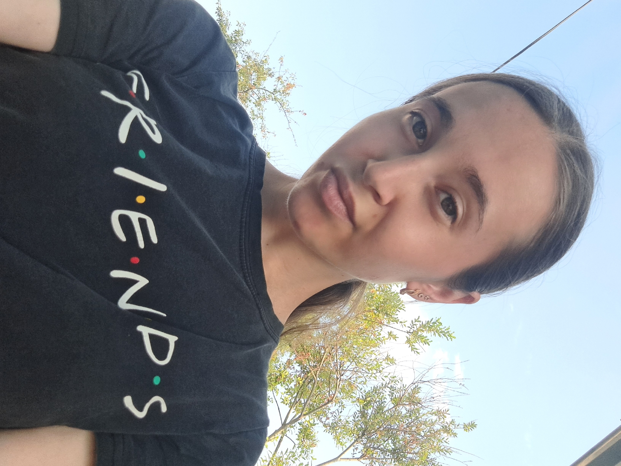 The image is a selfie of a young woman wearing a black t-shirt with the word "FRIENDS" printed on it in white and colorful dots. She has her hair pulled back in a ponytail and is wearing earrings. The background shows a blue sky with some trees. The image does not contain any specific details to indicate its location in the city of Vypasne, Ukraine. The scene is an outdoor setting, likely during the daytime, as indicated by the natural light and sky.