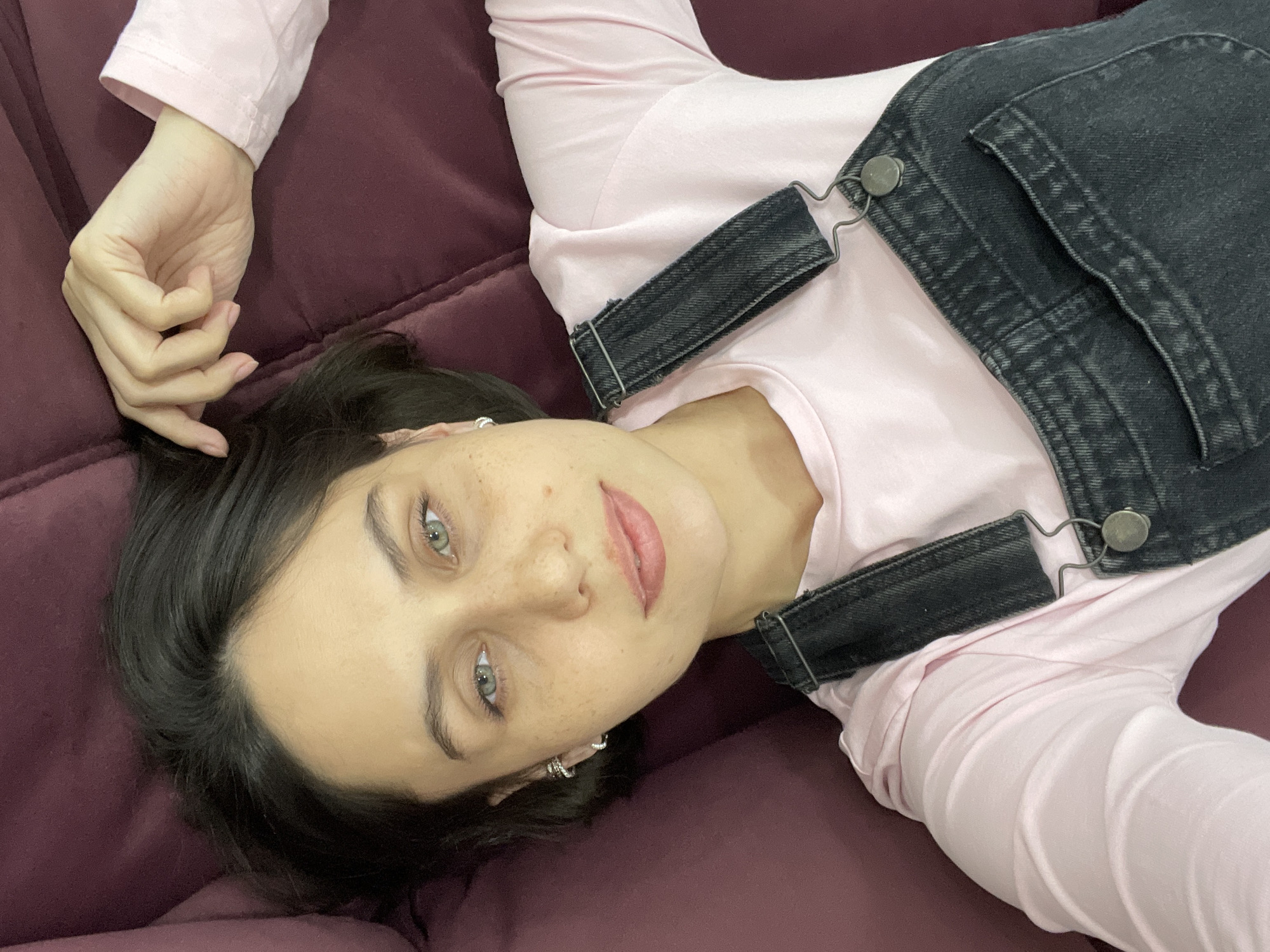 A young woman is depicted lying on a dark red or maroon sofa, looking directly at the camera. She has dark hair, styled with a side part, and striking green eyes. Her facial expression is neutral, with a slight, subtle smile on her tinted lips. Freckles are visible across her nose and cheeks. She is wearing a light pink long-sleeve top underneath dark grey or black denim overalls, which feature metallic buckles on the straps. Small, silver hoop earrings adorn her ears. Her left hand is visible near her head, with fingers gently curled. The lighting indicates an indoor setting, creating a relaxed atmosphere. There is no visible text in the image. This scene is set in Izhevsk, Russia.