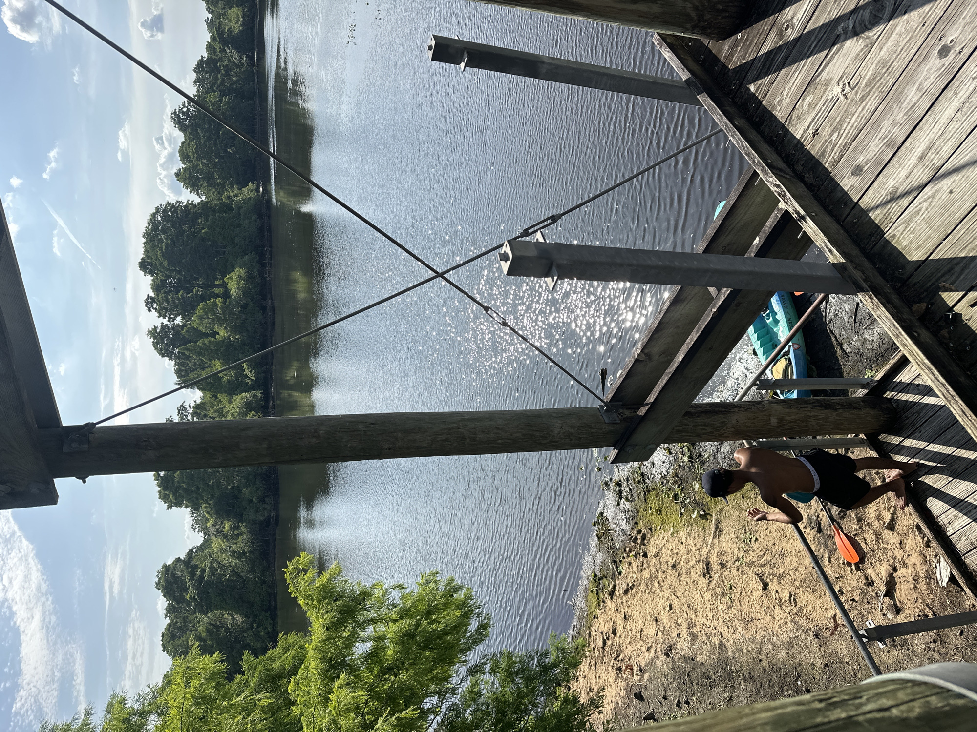 The image displays a sunny waterfront scene, likely a lake or wide river, viewed from an elevated wooden dock or pier. The setting is consistent with a recreational area in Tampa, United States.
In the foreground, beneath the dock, a young male is visible, shirtless and wearing dark swim shorts and a baseball cap. He is in a crouching position on a sandy and somewhat muddy bank. An orange and black paddle rests on the sand near him, and a blue and green kayak is partially visible, tucked close to the water's edge under the dock structure. He appears to be preparing for a water activity or exploring the shoreline.
The dock itself is constructed from weathered wooden planks and sturdy wooden beams, reinforced by diagonal metal supports and cables that crisscross the frame. Beyond the dock, the water surface is significantly rippled and sparkles with bright sun glare, indicating a clear, sunny day. Shadows from the dock stretch across the water and the sandy bank. The opposite shore is lined with dense, lush green trees, suggesting a natural and undeveloped landscape. Closer to the viewer on the lower left, vibrant green foliage from another tree is visible. The sky above is a bright blue with scattered white clouds, one showing a faint contrail. No discernible text is present in the image.