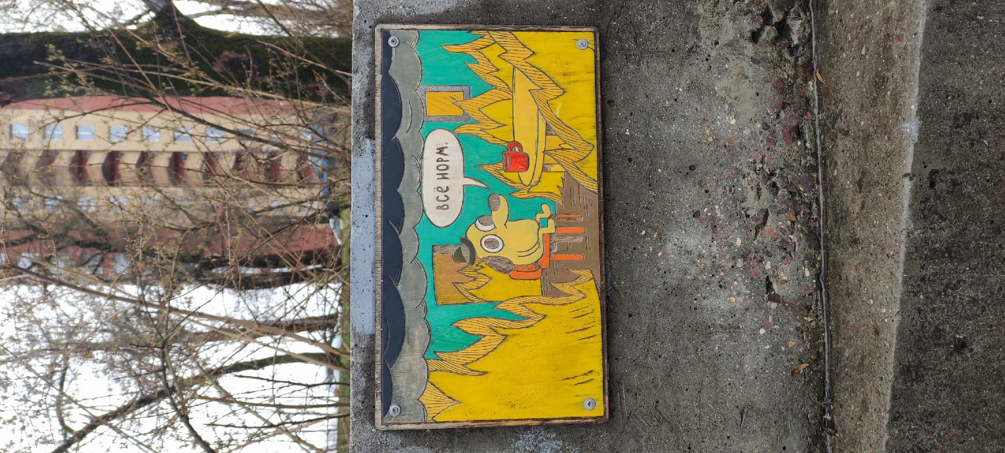 A colorfully painted sign is affixed to a gray concrete wall or pillar. The sign depicts a yellow cartoon figure with large eyes, seated in a chair, with a red cup nearby. A speech bubble contains text in Russian, "ВСЁ НОРМ." The background behind the figure is divided into turquoise and yellow sections, with stylized depictions of trees or foliage. Fasteners are visible in the four corners of the sign. The sign's border appears to be made of wood. Outside of the wall, a tree is seen with bare branches, and a building is slightly out of focus in the background. The scene is located in Shchyolkovo, Russia.