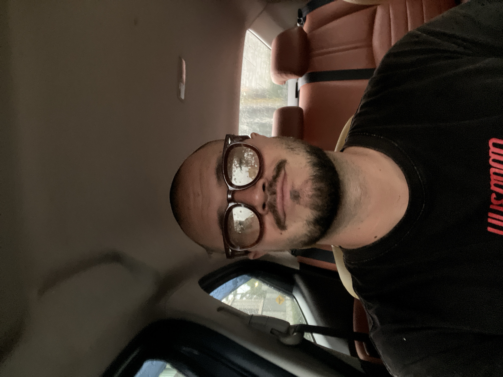 A man is captured in a selfie taken from inside a car. He is seated in the back seat, facing the camera. The man has a shaved head, a mustache, and a goatee. He is wearing thick-rimmed glasses and a black t-shirt. The interior of the car is visible, with brown leather seats and the ceiling of the car above. Through the window behind him, the weather appears to be rainy, with blurred trees and buildings visible in the distance. The overall lighting suggests it is either overcast or nighttime.