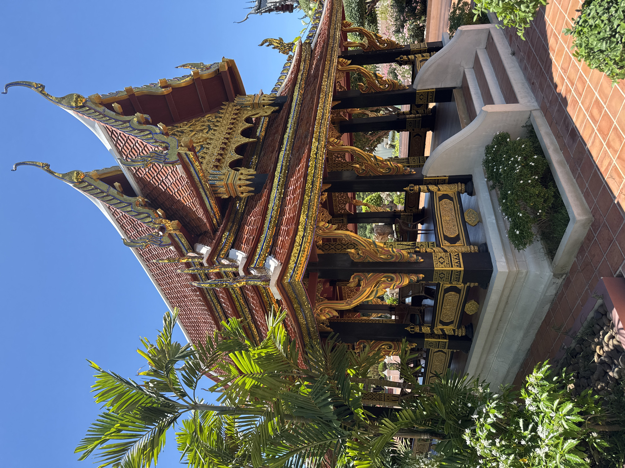 The image depicts a traditional Thai building located in the Bang Pu Subdistrict Municipality, Thailand. The building features a multi-tiered, red-tiled roof with elaborate gold ornamentation along the edges and gables. Black pillars adorned with golden carvings support the structure. A set of stairs leads up to a platform adjacent to the building, decorated with white concrete and flower beds with green plants. A brick-paved area surrounds the building's base. Lush green foliage, including palm trees, is visible in the foreground. The sky is a clear blue.
