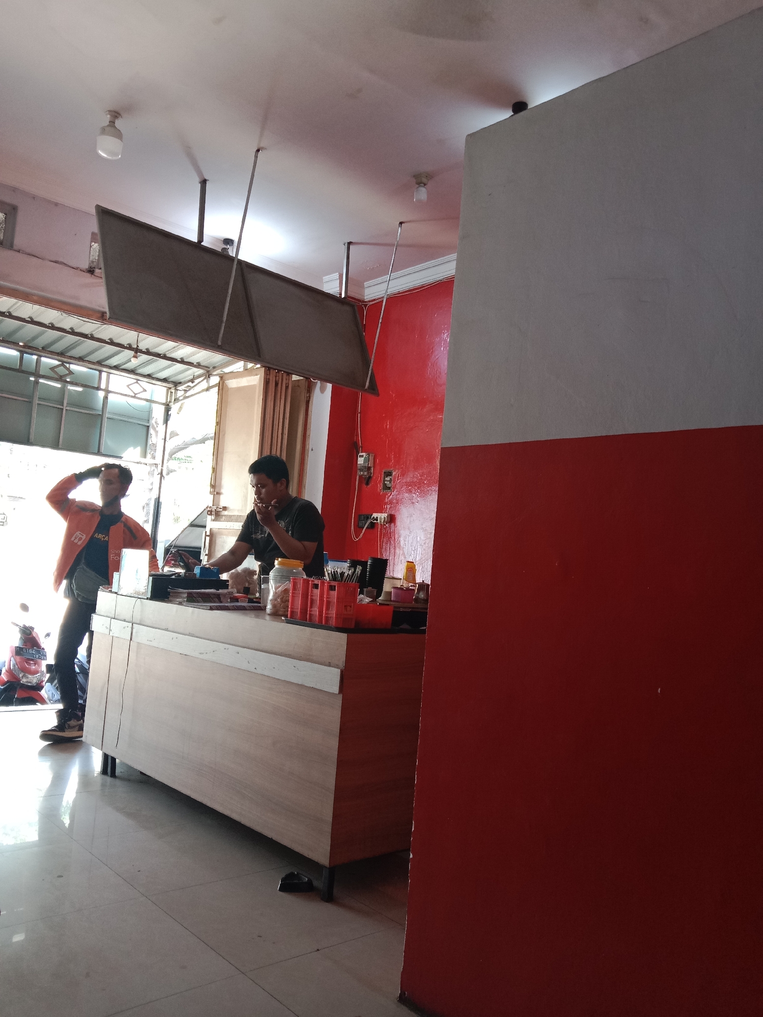 The image depicts the interior of a small establishment, likely a food stall or shop, located in Kaliwungu, Indonesia. Two men are visible. One man is wearing an orange jacket with the word "ARCA" printed on it, indicating he may be a delivery driver. He is standing near the front, holding his head. The other man, wearing a black t-shirt, is behind a wooden counter, eating. The counter is cluttered with various items including containers, utensils, and possibly food products. A sign is suspended above the counter. Behind the counter, the wall is painted red. The foreground is dominated by a large, wall painted with a gray upper section and a red lower section. A red scooter is parked outside the establishment. The lighting is somewhat dim, with natural light coming from the outside. The floor is tiled.