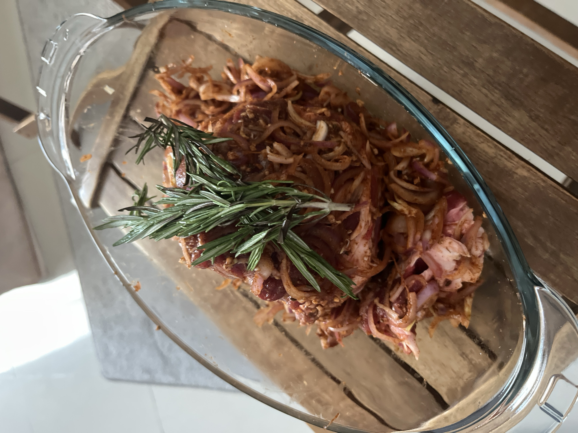 A close-up, overhead shot shows a glass baking dish filled with marinated meat and sliced onions, topped with a sprig of rosemary. The dish is placed on a wooden table, with slats visible. The lighting suggests an indoor setting with natural light coming from the left. The scene appears to be a kitchen or dining area, possibly in Dubai, United Arab Emirates, given the context of the prompt. There are no people or animals visible in the image, and the focus is entirely on the food preparation.