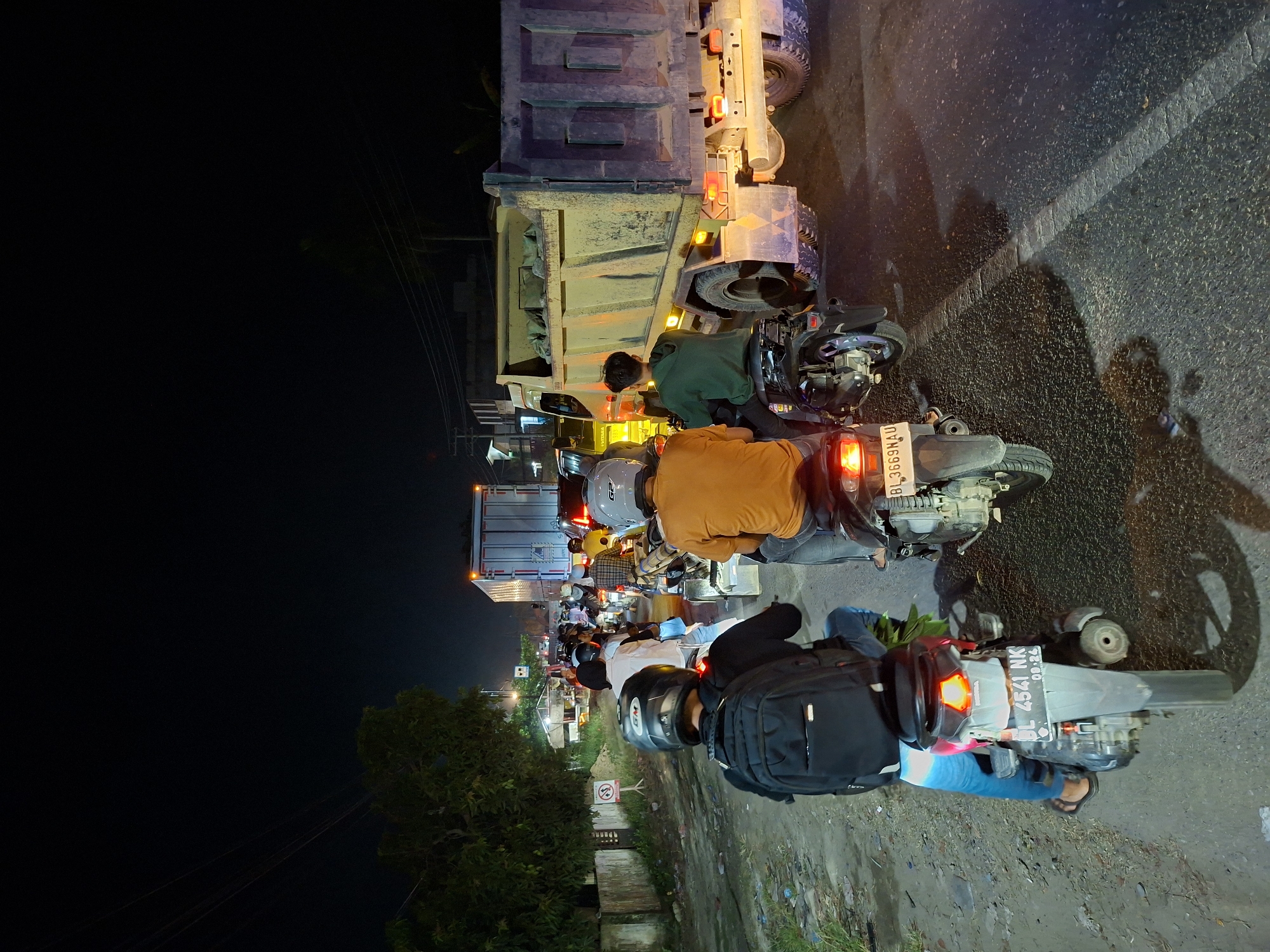 The image captures a nighttime scene of slow-moving or stationary traffic in Lhokseumawe, Indonesia, viewed from behind a motorcyclist. The perspective is vertically oriented, looking forward along a road illuminated primarily by vehicle lights.
In the immediate foreground, the back of a person wearing a black backpack and a helmet is visible, sitting on a motorbike. The motorbike has a license plate "BL 4541 NK" with "08.24" below it. To the right, another person on a motorbike, wearing an orange shirt and a light-colored helmet, is visible, with their motorbike's taillight illuminated. Its license plate, "BL 3669 AU", is partially visible. Further ahead, another individual in a green hoodie is positioned near the front of a large, yellow dump truck.
The dump truck, bearing the partially visible brand "HYUNDAI", is a dominant feature in the midground, its various amber lights illuminated. Behind it, a queue of other motorbikes and at least one more truck or larger vehicle stretches into the distance, their headlights and taillights creating streaks of light against the dark background. The road surface is asphalt, with a white line marking the edge or center. A dirt shoulder runs along the right side of the road, showing some debris and puddles.
To the left, beyond the traffic, the scene darkens considerably. Silhouettes of trees are visible, along with power lines crisscrossing the dark sky. A small, white and red circular sign, possibly indicating 'no entry' or a similar prohibition, stands on the left side of the road. Buildings with some illumination can be faintly discerned in the far background. The overall impression is one of a busy urban or semi-urban road at night, characterized by vehicular congestion.