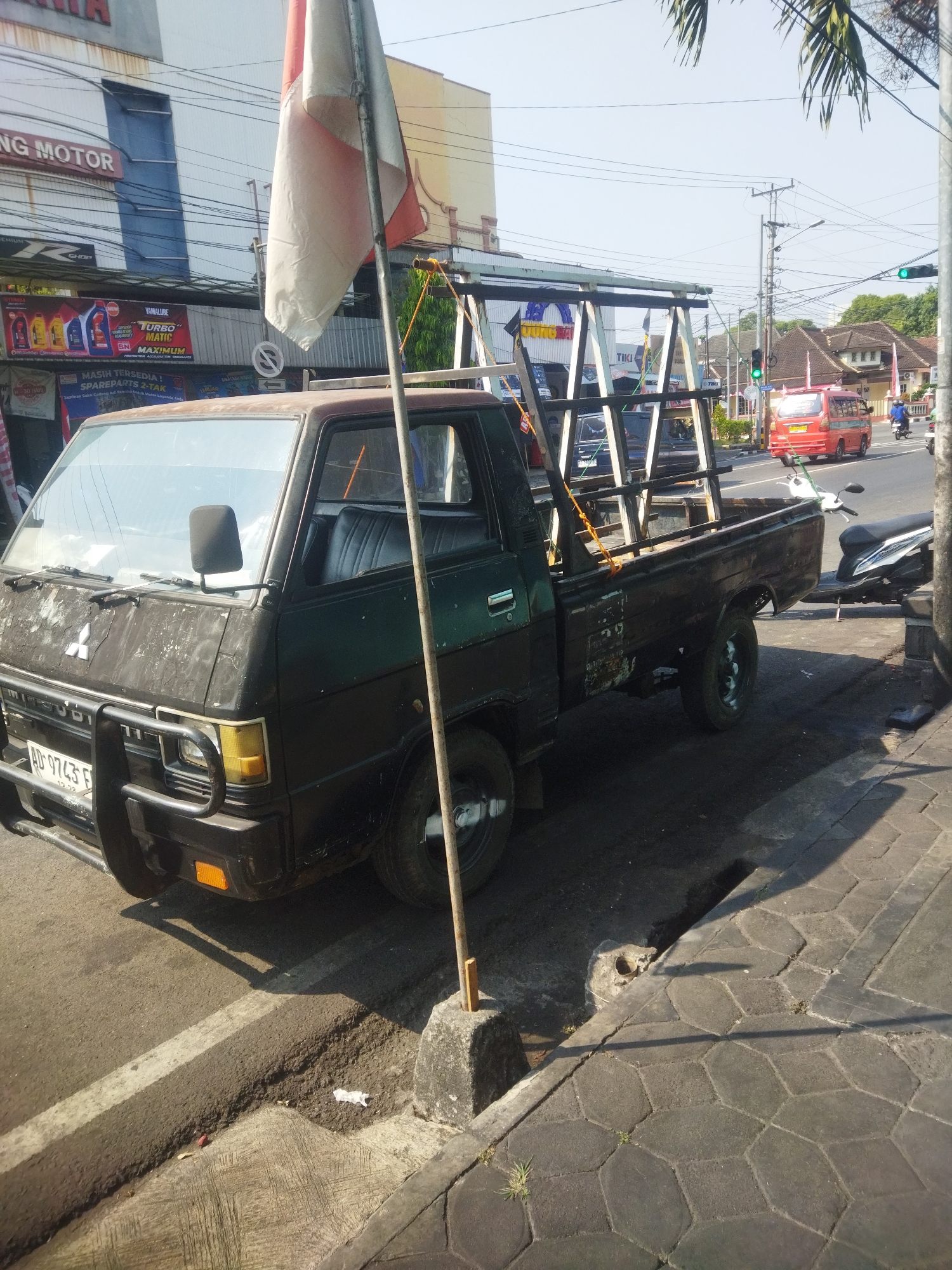 A black Mitsubishi pickup truck with a faded exterior is parked on the side of a street in Giripurwo, Indonesia. The truck is adorned with a weathered Indonesian flag attached to a metal pole. In the background, a red van, a motorcycle, and various buildings are visible. The street features a white line marking the edge of the road, and a sidewalk paved with hexagonal stones. The scene is illuminated by sunlight, casting shadows on the ground.