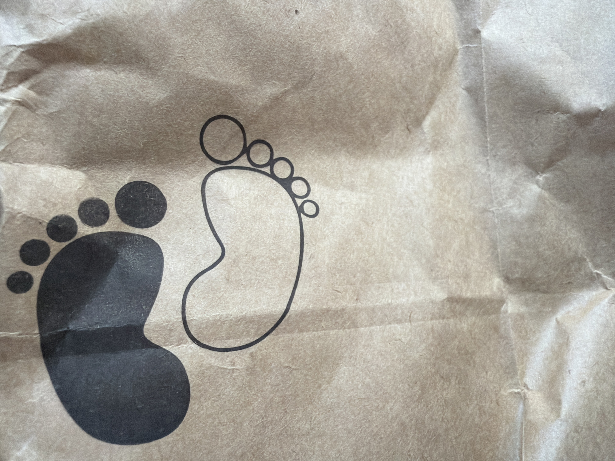 The image displays a close-up of a brown paper bag with black ink designs of two feet. One foot is a solid, large imprint, while the other is an outline with circles representing toes. The paper bag is crumpled, showing folds and creases. There are no people or any indications of the city of Qarağandı city administration, Kazakhstan. The image is purely focused on the paper bag's design and texture.