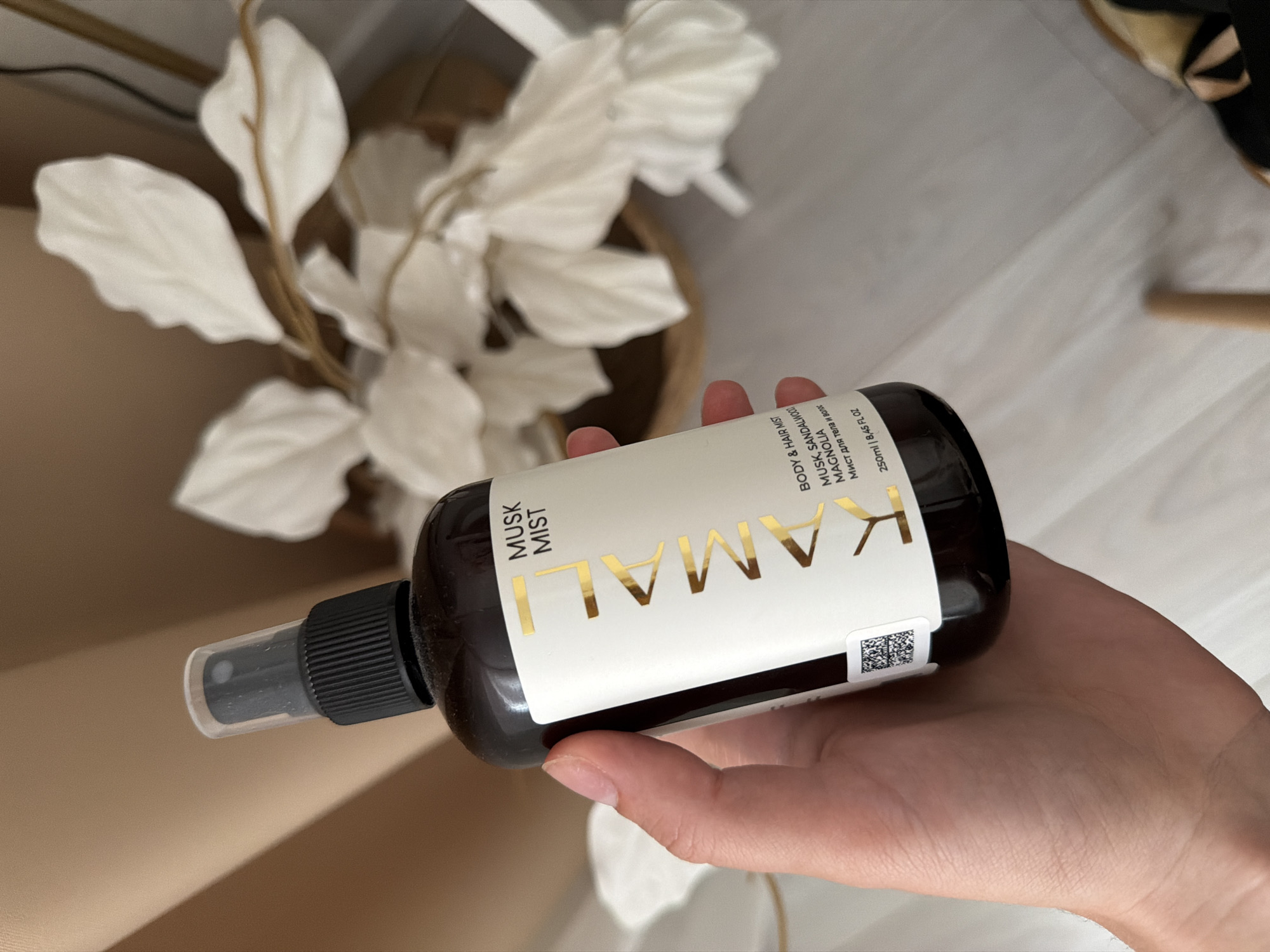 In this close-up shot, a person's hand holds a dark brown spray bottle labeled "KAMALI MUSK MIST." The bottle has a white label with gold lettering, featuring the brand name "KAMALI" prominently. Below the brand name, the label indicates "BODY & HAIR MIST" and lists the scent notes as "MUSK, SANDALWOOD, MAGNOLIA." There's also text in Cyrillic, "Мист для тела и волос," confirming the product's purpose. The bottle contains 250ml or 8.45 fl oz. The background is softly blurred, showing muted tones of beige and white, with a hint of what appears to be dried decorative branches or flowers in a basket, suggesting an indoor setting with a focus on aesthetics and perhaps self-care products. The lighting is soft, possibly from natural daylight, creating a gentle and inviting atmosphere. The image focuses on the product being held, with the hand gently supporting the bottle, implying a sense of care and presentation.