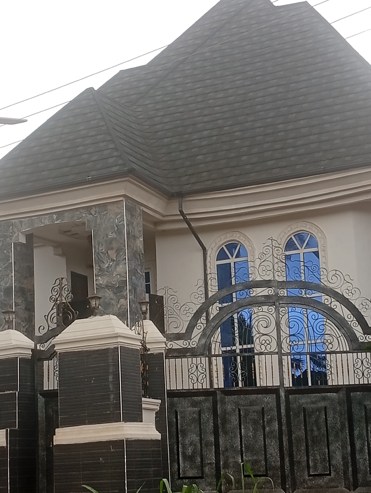 The image shows the exterior of a house in Akokwa, Nigeria. The house features a dark grey, textured roof, large arched windows with blue glass and white frames, and decorative iron gates. The entrance has a covered porch supported by columns clad in grey and white marbled material. The gate posts are black and white, and there are decorative iron elements on the gate. The scene appears to be captured during the day, with overcast sky conditions, suggesting a cloudy or hazy atmosphere. The foreground shows some greenery, likely plants or grass.