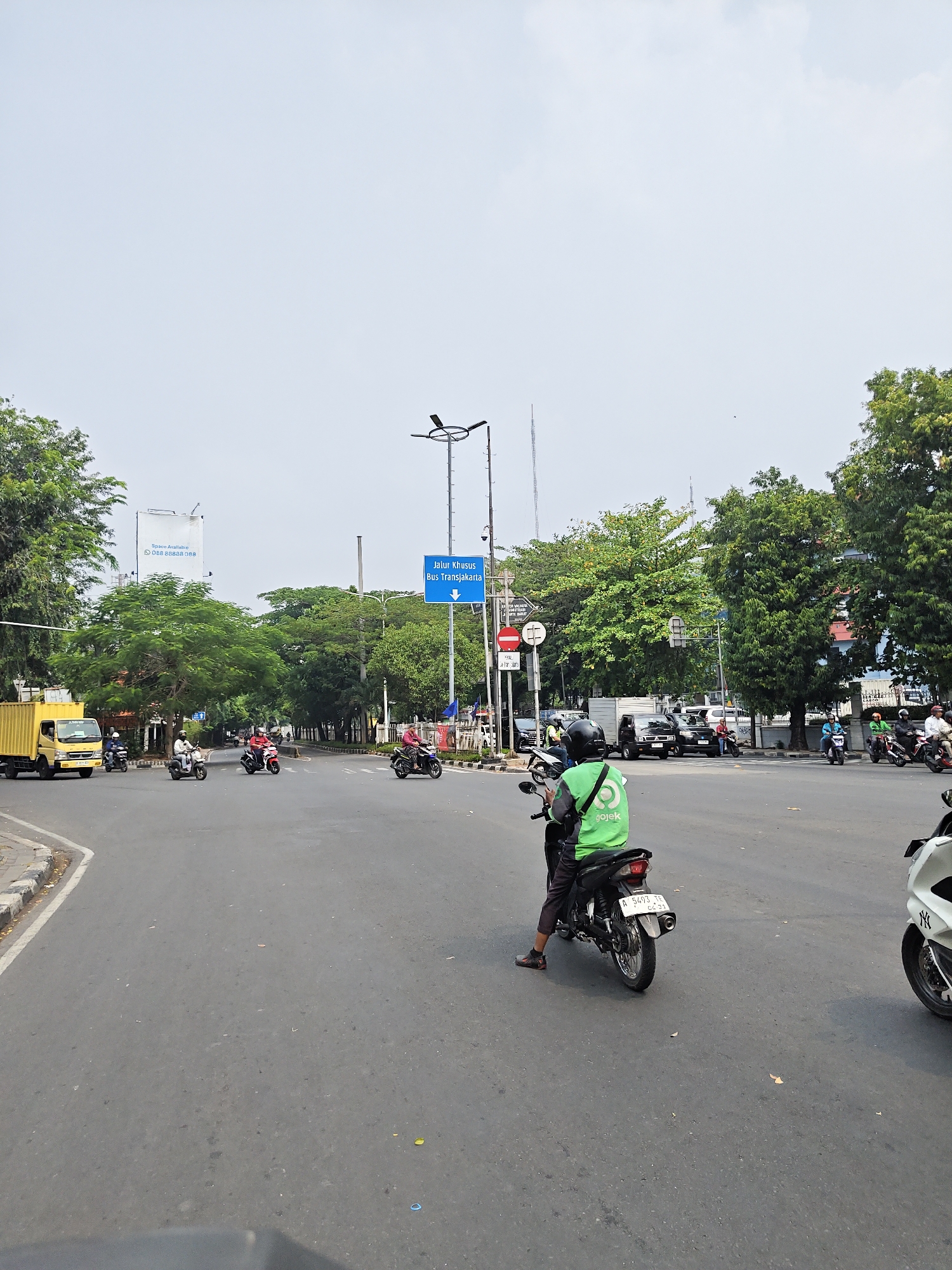 The image shows a street scene in West Jakarta, Indonesia, likely a busy intersection. A yellow truck is on the left, and several motorcycles are visible. One motorcycle in the foreground has a rider wearing a green vest with the "Gojek" logo. The road is paved, and there are trees and buildings along the sides. A blue sign indicating "Jalur Khusus Bus Transjakarta" (Dedicated Bus Transjakarta Lane) is prominent. The sky is overcast.