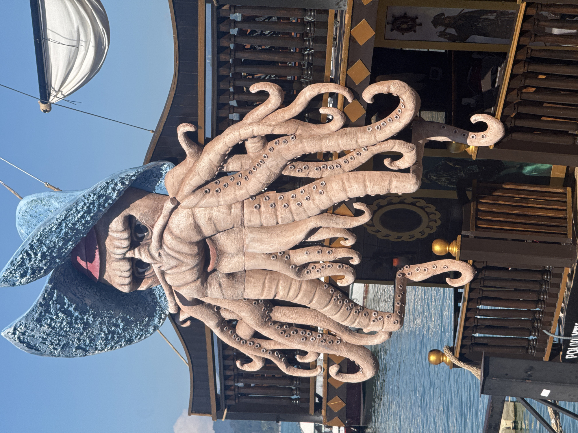 The image depicts a maritime-themed structure, possibly a restaurant or entertainment venue, located in Batumi, Georgia. The primary feature is a large, sculptural representation of an octopus wearing a pirate hat. The octopus, rendered in a tan color with numerous suckers visible on its tentacles, is mounted on the side of the building. The building itself has a dark brown wooden structure with gold accents, resembling the architecture of a pirate ship. A partial view of a white sail is visible in the upper left corner of the image. The sky is clear and blue. Body of water is in the background.