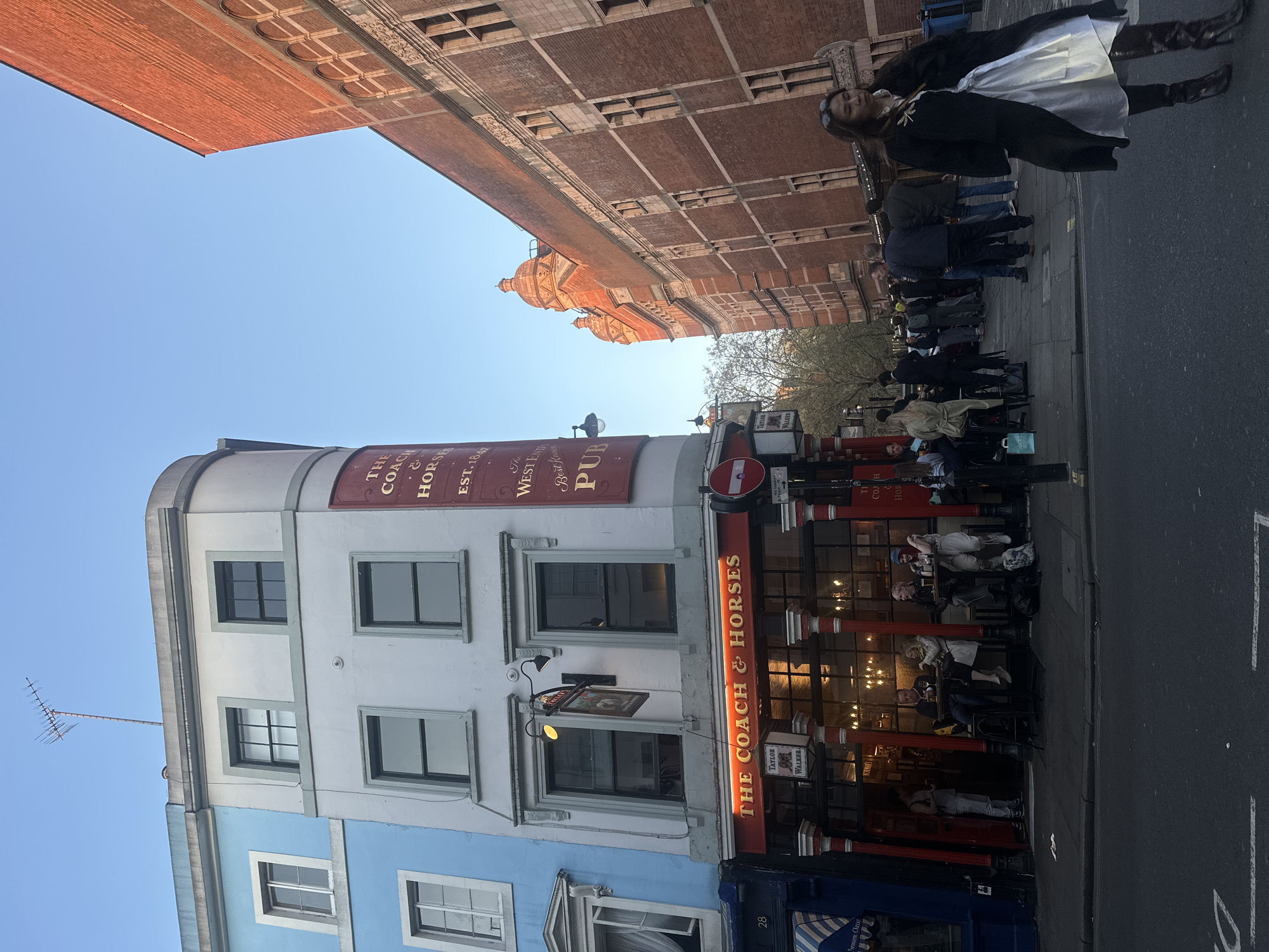 The image shows The Coach & Horses pub in Soho, London. The pub's exterior is painted white and blue, with red signage. A red sign above the entrance says "THE COACH & HORSES." People are seated at tables outside the pub. A woman in a black coat and white skirt is walking on the sidewalk. Buildings are visible in the background, including one with a brick facade. The sky is blue.