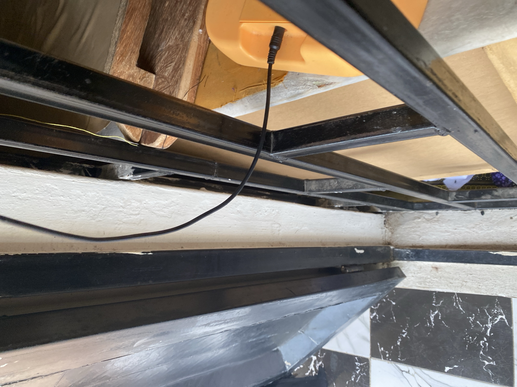 The image shows a close-up view of a black metal gate or security bar system, possibly in a doorway. The gate is partially open, revealing a white wall and a tiled floor with a black and white marble pattern. A black electrical cable runs from the left of the frame towards a bright orange object on the right, which appears to be a power adapter or a similar electronic device. A thin yellow wire is also visible behind the metal bars. The scene suggests an outdoor or semi-outdoor environment, potentially a residential or commercial property in Jalingo, Nigeria. The lighting is natural, indicating daytime. There are no people visible in the image.