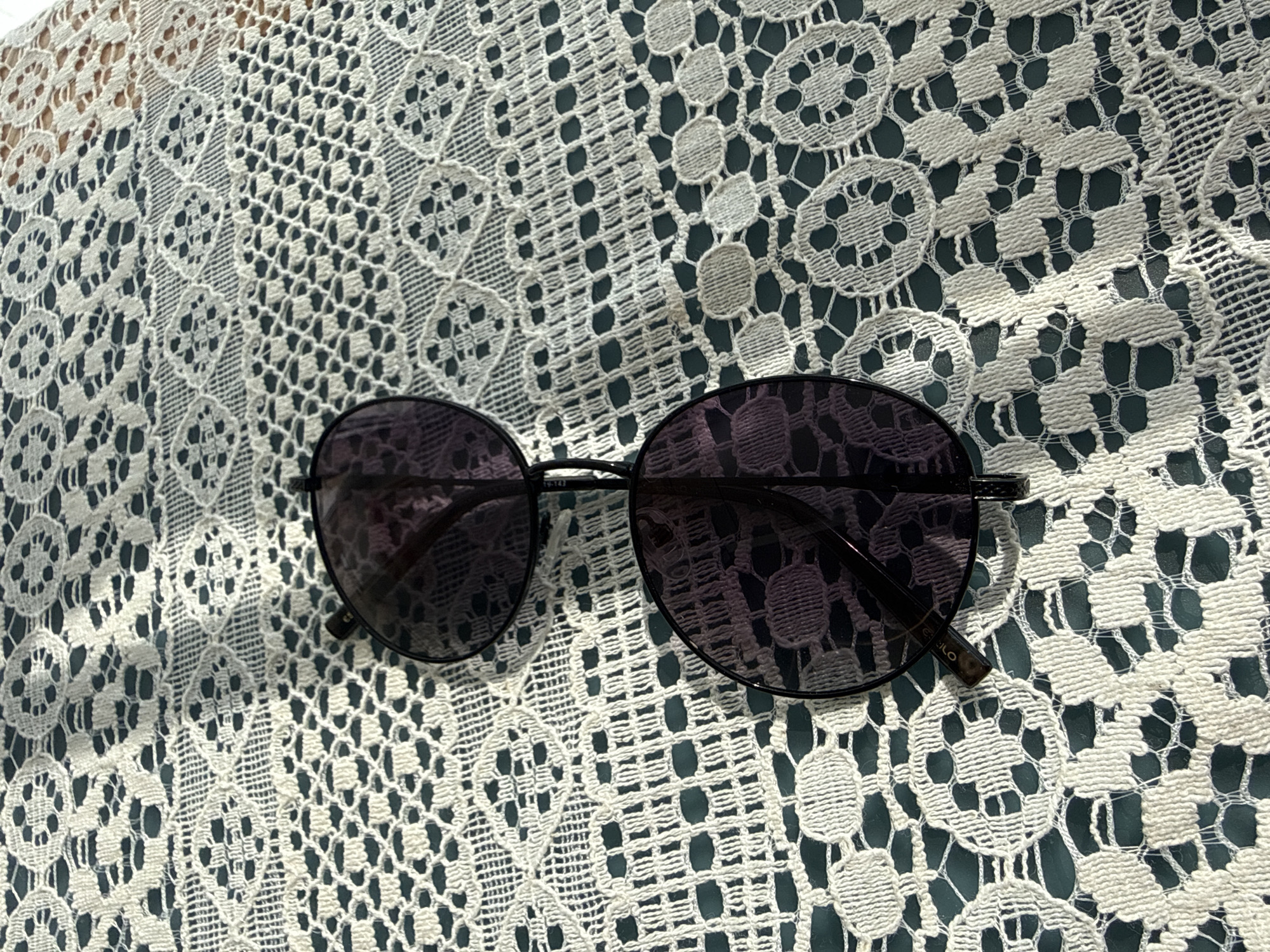 A pair of dark-lensed sunglasses rests on a white, intricate lace tablecloth with a dark green background. The sunglasses are positioned diagonally across the frame, with the lenses facing upwards. The lace pattern is detailed, featuring circular motifs and geometric designs. The image appears to be taken indoors or in a shaded outdoor area, given the lighting conditions and the absence of strong shadows. There are no people or any other objects visible in the frame. The scene lacks any identifiable landmarks or context that would suggest a specific location within Sochi, Russia, or any other city.