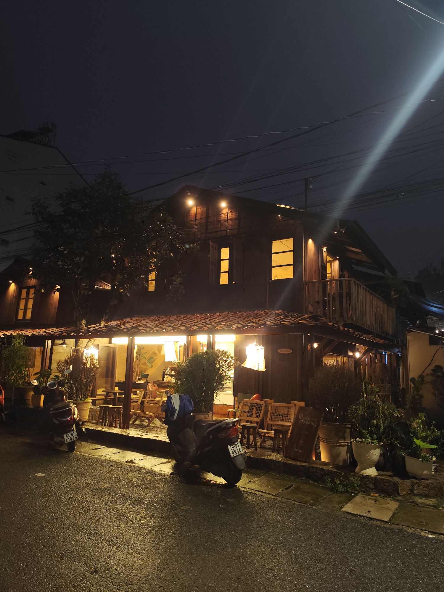 A two-story wooden building with a red tile roof, likely a cafe or restaurant, is illuminated at night on a wet street in Da Lat, Vietnam. The building has multiple windows, some of which glow with warm, yellow light, suggesting an interior space. A balcony with a wooden railing extends from the upper floor. The ground floor features an open-air seating area with wooden chairs and tables, and several potted plants. Two motorbikes are parked on the street in front of the building. The street is paved and appears to be made of asphalt, with a sidewalk or raised area leading to the building's entrance. Overhead, power lines crisscross the dark night sky.