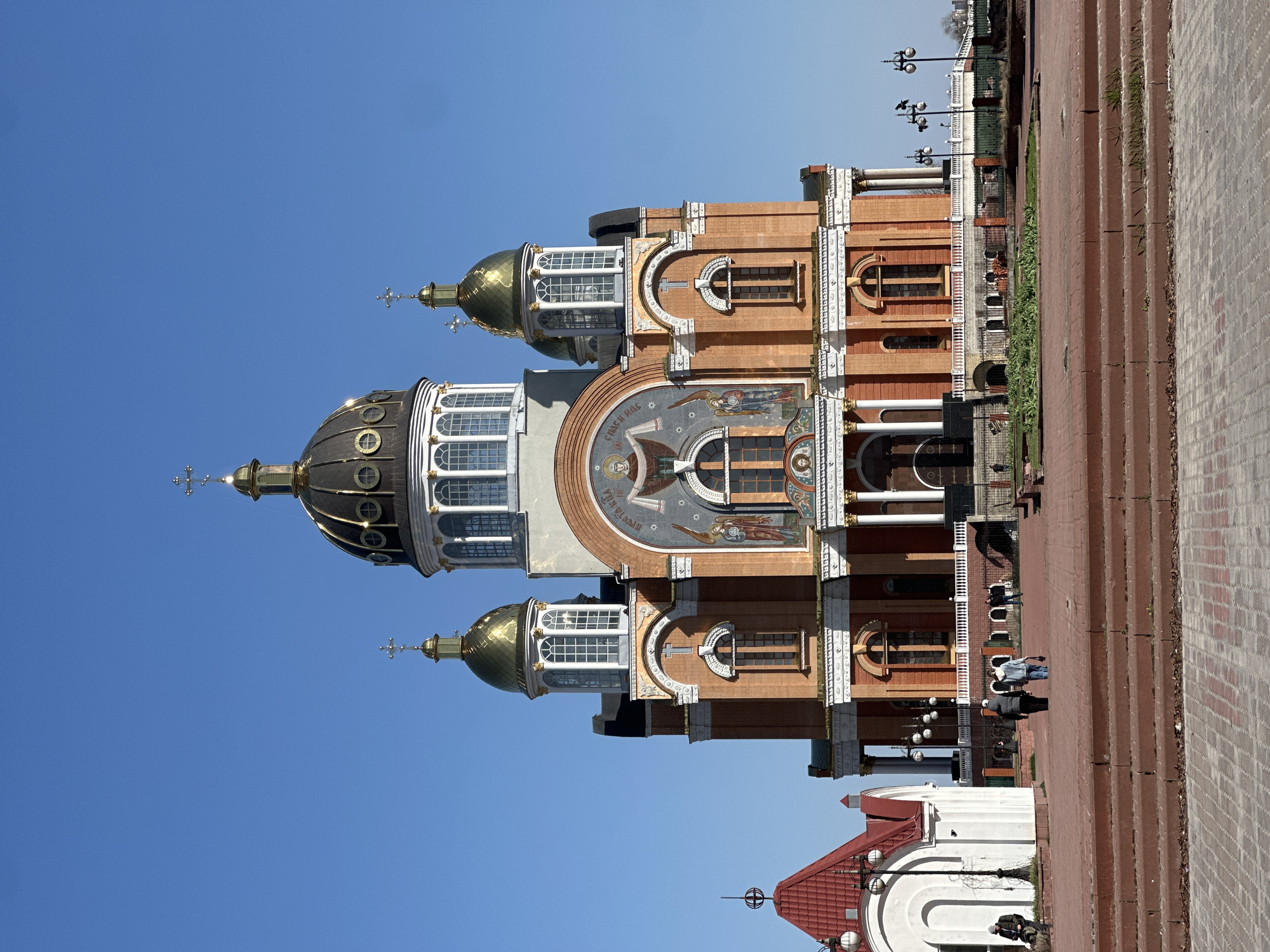 The image is of the Ukrainian Orthodox Church of Moscow Patriarchate Cathedral of the Resurrection of Christ in Kyiv, Ukraine. The cathedral has multiple golden domes and is made of red brick. There are people walking on the red brick pedestrian walkway in front of the church. The sky is blue.