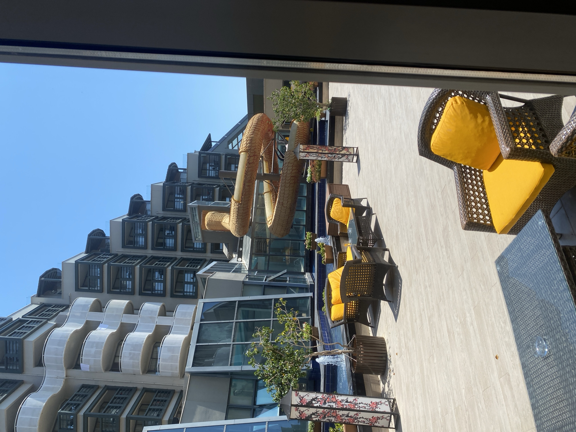 This is a photograph of an outdoor patio area adjacent to a resort or hotel complex in Aksu, Turkey. The scene depicts a water slide in the shape of a coiled serpent, a swimming pool, and an outdoor seating area with wicker furniture and bright yellow cushions. The buildings in the background are modern and multi-storied, with balconies and large glass windows. The sky is clear and blue, suggesting a sunny day. The patio floor is paved with light-colored tiles. The overall impression is that of a resort amenities area designed for relaxation and recreation.