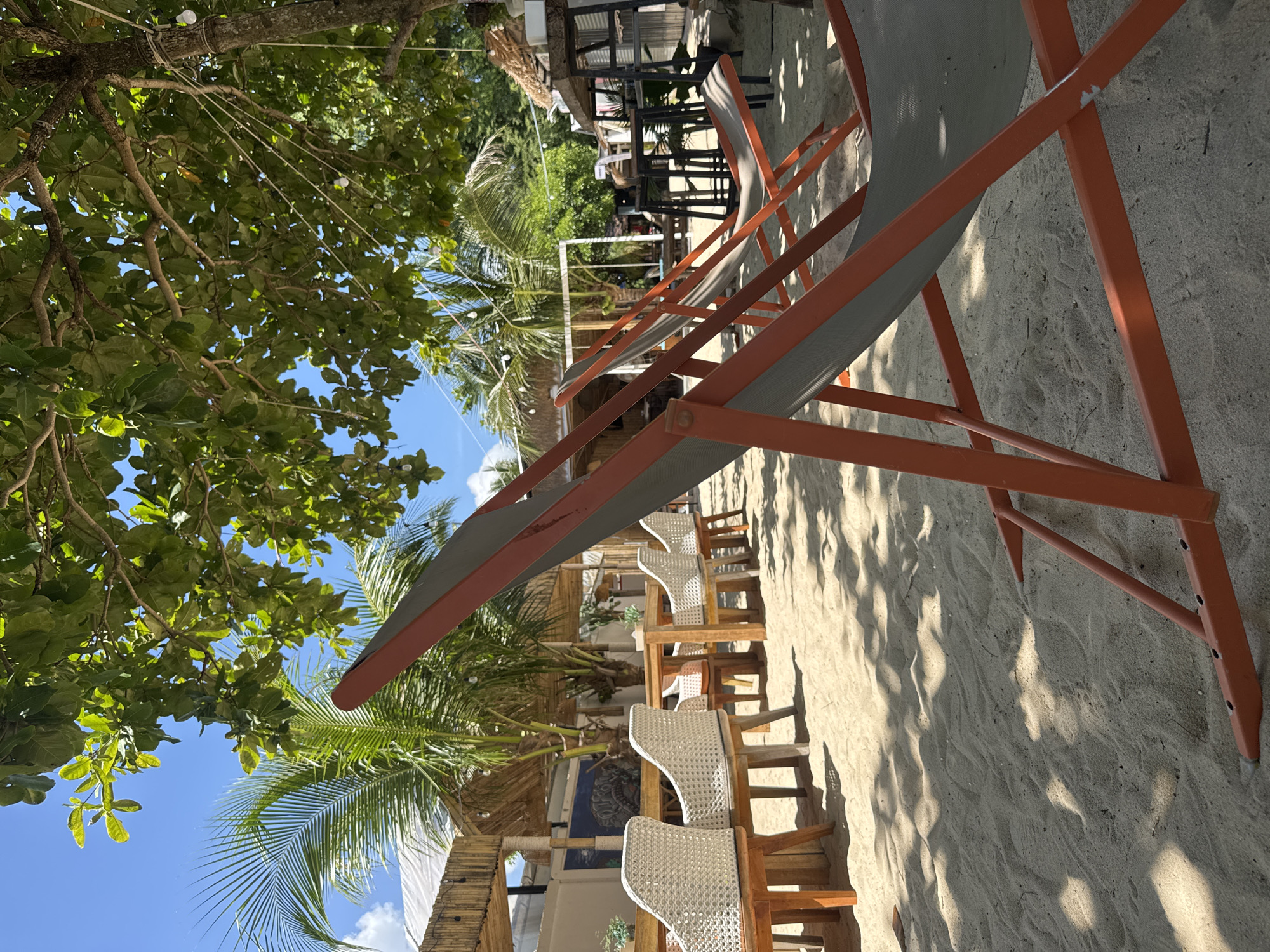 The image shows an outdoor seating area on a sandy beach, likely in Choeng Thale, Thailand, with a tropical ambiance. In the foreground, two orange-framed beach chairs with grey fabric are set up on the sand, casting shadows from the overhead tree foliage. Behind the chairs, several wooden tables and white woven chairs are arranged on the sand, suggesting a dining or lounging area. In the background, there are thatched-roof structures that appear to be part of a restaurant or bar, with palm trees and lush green foliage contributing to the tropical setting. The sky is bright blue with scattered clouds, indicating a sunny day. The overall scene depicts a relaxed, beachside establishment designed for patrons to enjoy the coastal environment. There are no people visible in the image.