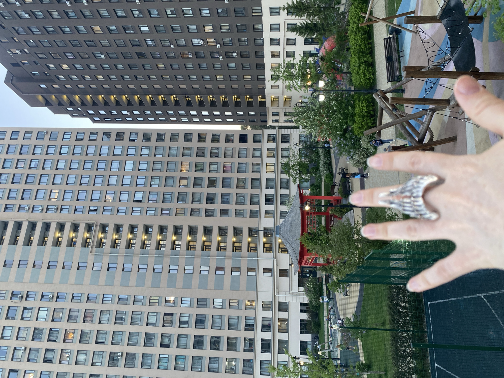 The image shows a residential area in Yekaterinburg, Russia, with two tall apartment buildings in the background. The buildings are composed of multiple floors with many windows. Between the buildings, there is a park with a red pagoda structure. A playground with wooden structures and a black net is also visible in the park. In the foreground, a hand with a large, silvery ring is outstretched towards the scene. There are trees and a green fence in the midground.