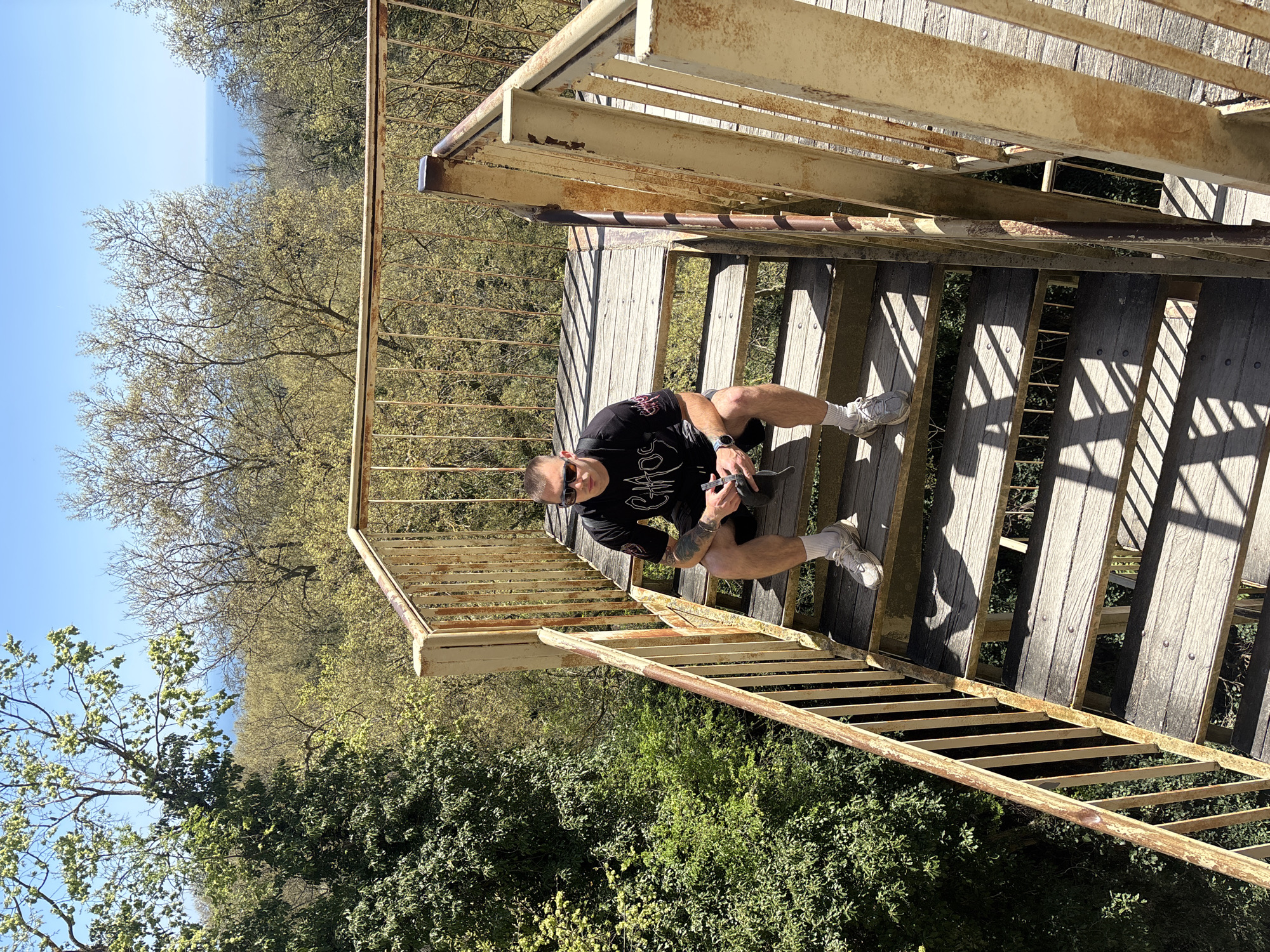 The image shows a man sitting on wooden stairs with a metal railing. The man is wearing sunglasses, a black t-shirt with a graphic design, and shorts. He is holding a small object in his hands. The stairs are surrounded by green trees and foliage. In the distance, there is a body of water. The location is in the city of Unknown, Unknown.
