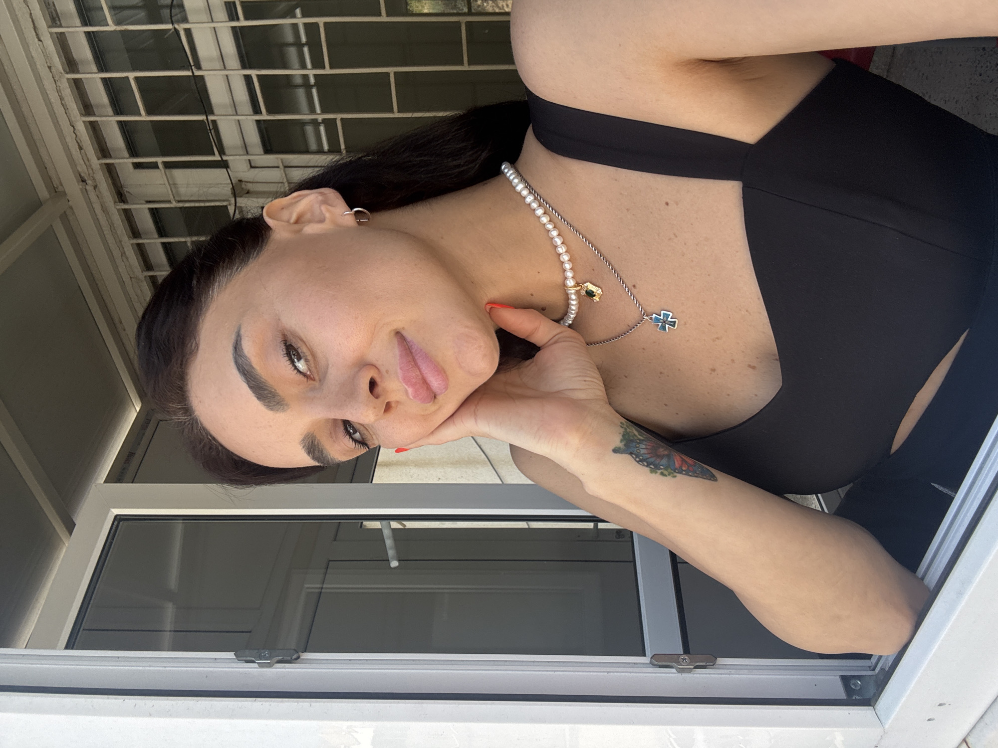 A young woman with dark hair, a pale complexion, and dark eyebrows is pictured from the chest up, looking towards the viewer. She is leaning her left hand against her cheek and touching her face with her fingers. She is wearing a black top with thin straps and a cutout design, a pearl necklace with a gold pendant, and a silver chain with a cross pendant. She has a tattoo of a butterfly on her left forearm. She is positioned near a white window frame. The background shows a building with barred windows, suggesting an urban environment. The lighting indicates it is daytime, and the scene appears to be outdoors or in a well-lit indoor space. The setting does not provide specific details to pinpoint the location to Tbilisi, Georgia.