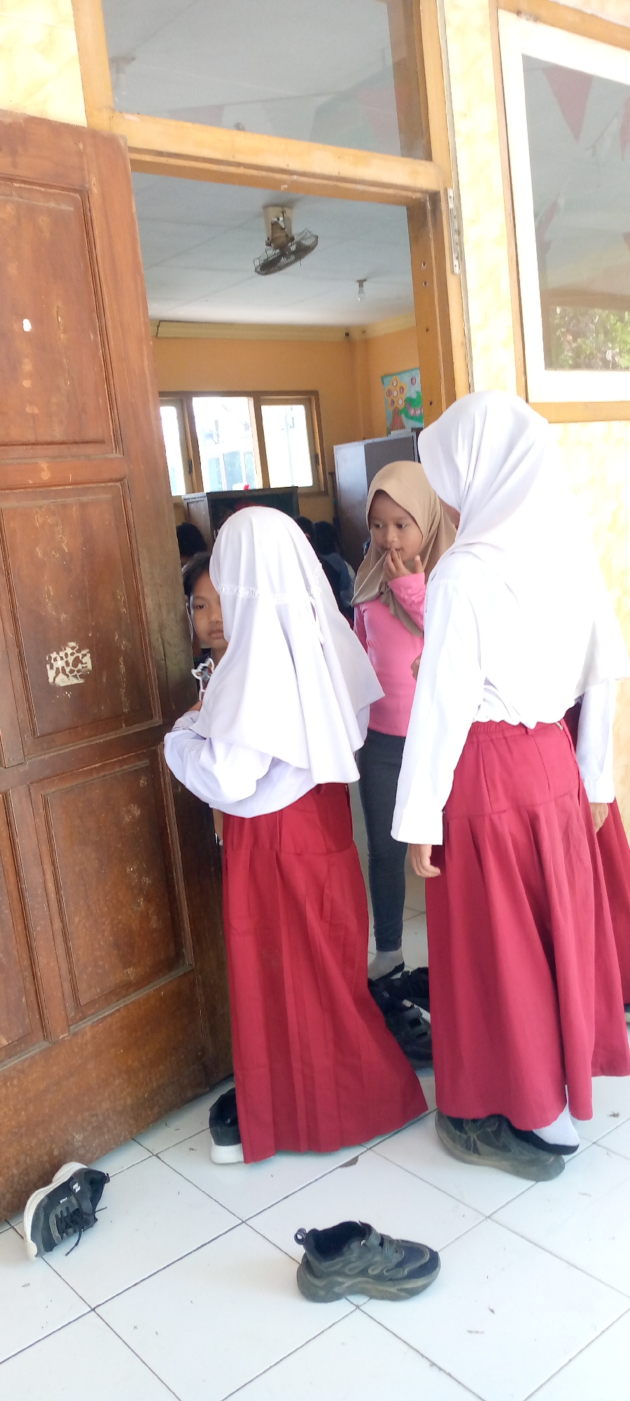 In a hallway with a tiled floor and cream-colored walls, three young girls are visible. Two girls are wearing white hijabs and long red skirts. One of these girls is partially obscured by a wooden door. The third girl is wearing a beige hijab and a pink long-sleeved shirt, with dark pants. She is looking to her left and has her hand on her chin. In the background, a ceiling fan is visible, and a framed picture hangs on the wall. Several pairs of shoes are on the floor. The location is identified as Pacing, Indonesia.