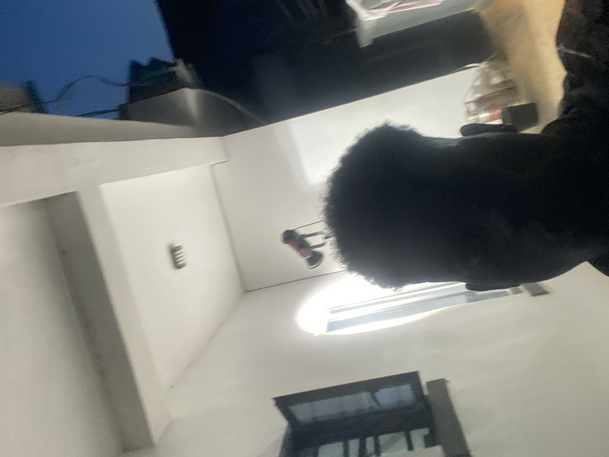 The image shows an indoor scene with a person in the foreground, their head and shoulder visible. The environment appears to be a room with white walls. A window is visible towards the bottom of the frame. A ceiling fan and other unidentified objects are present on the ceiling. The location is unknown.