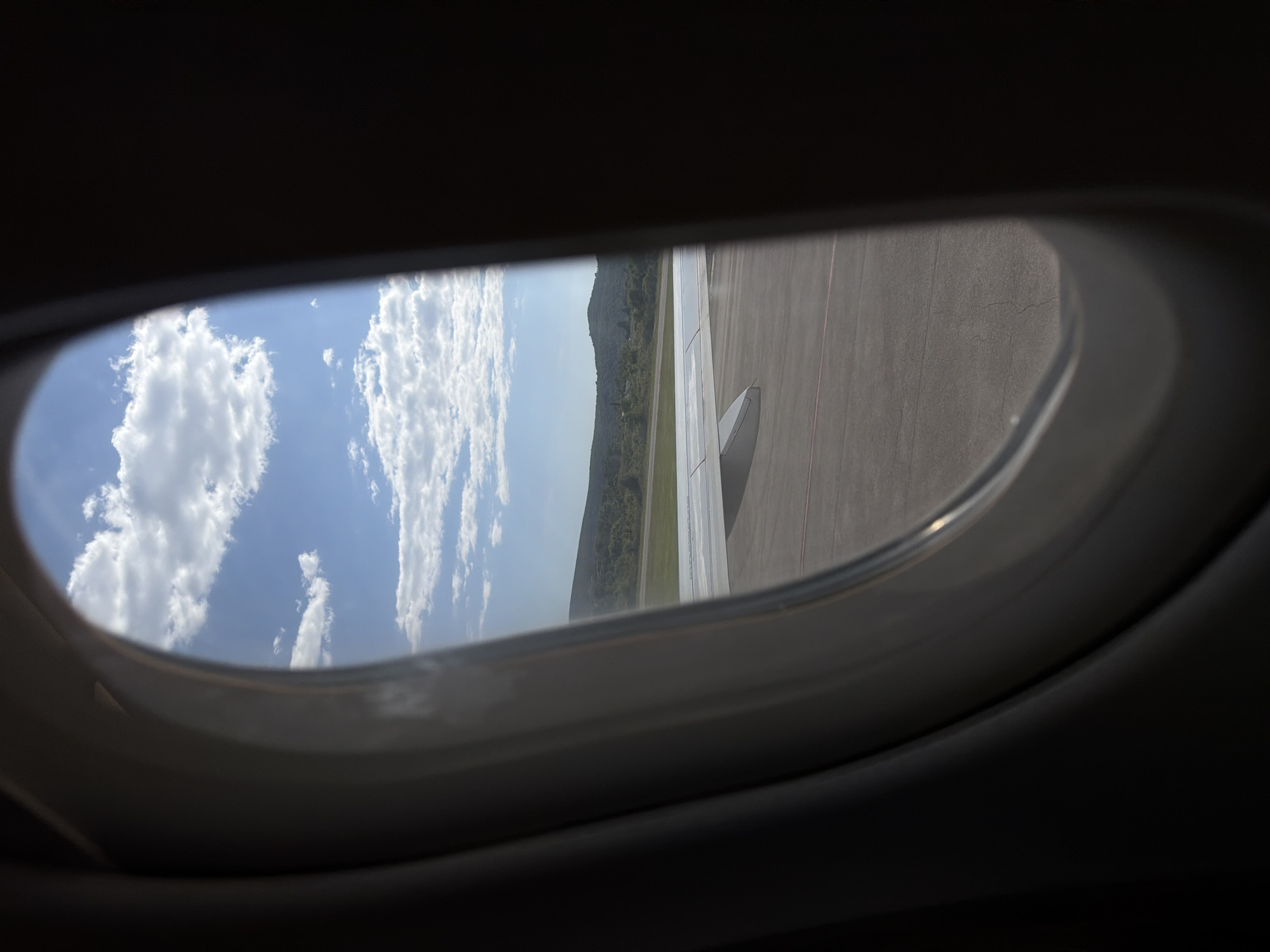 The image is taken from inside an airplane looking out a window. Outside the window is a view of the wing of the plane, an airport runway, a grassy area, and a forested hillside against a backdrop of a blue sky with white clouds. The scene appears to be during daylight hours. The location is uncertain without additional context.