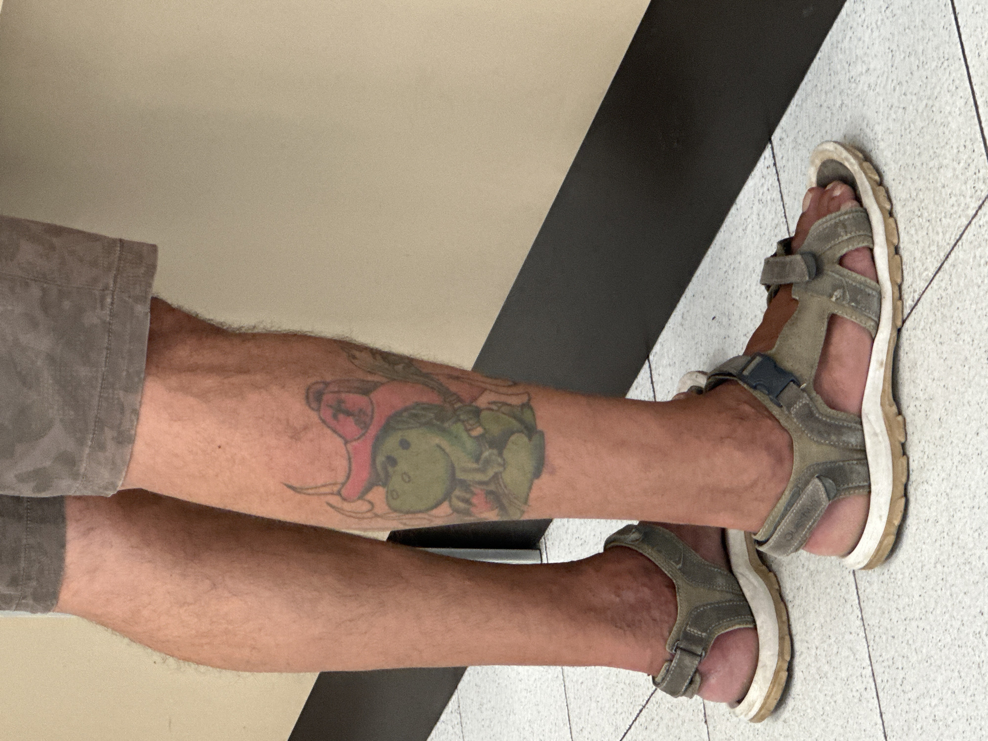 The image shows a close-up view of a person's legs and feet, wearing grey sandals. The person has a tattoo of a cartoon dinosaur wearing a red hat on their right calf. The background consists of a tiled floor and a wall with a black stripe. The scene is indoors, with the lighting suggesting an artificial light source. There is no visual information in the image that could place the scene in Trieste, Italy.
