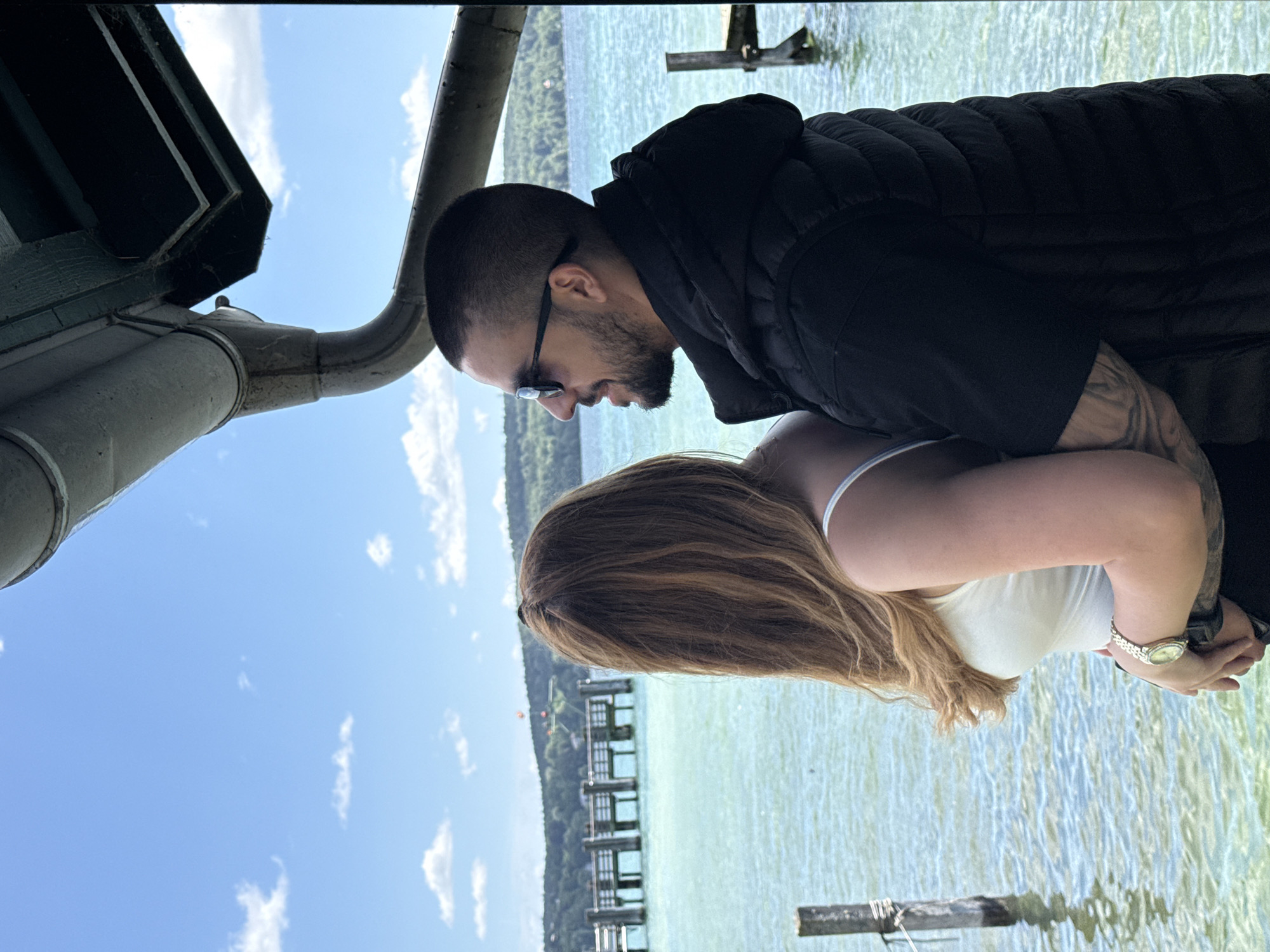 A man and a woman are standing on a pier, embracing, and looking out at a body of water. The man, on the right, is wearing a black puffer vest over a black jacket and sunglasses. He has dark hair and a beard. The woman, on the left, has long blonde hair and is wearing a white tank top. She has a watch on her left wrist. The water is a clear turquoise color. In the background, there is a wooden pier with railings and trees on the distant shore. The sky is blue with scattered white clouds. The location appears to be a lakeside setting, possibly in Starnberg, Germany, given the presence of a pier and a lake.
